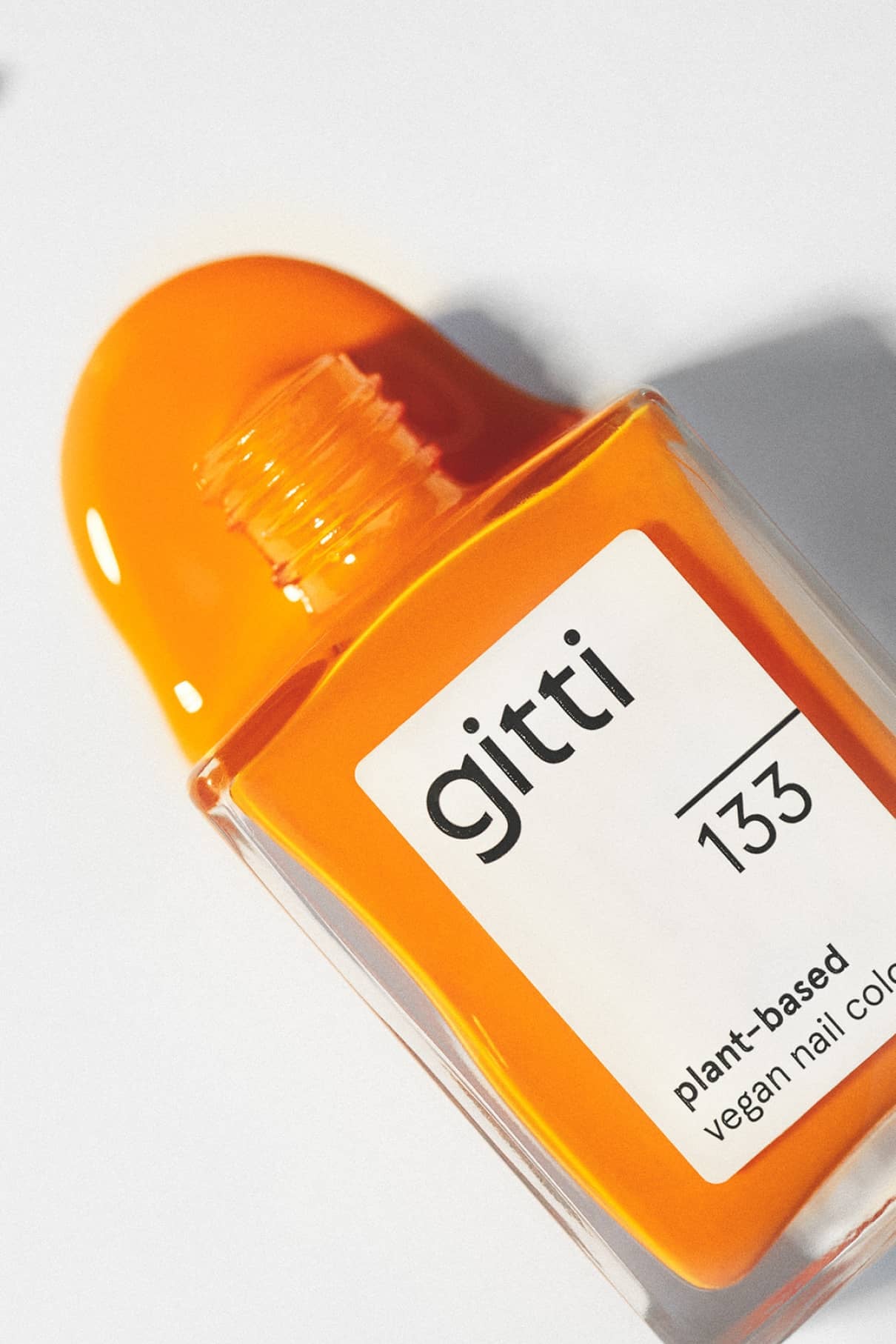 Eine offene Flasche gitti no. 133 vegane Nagelfarbe in leuchtendem Orange steht auf einer weißen Fläche, daneben ist etwas Lack verschüttet.