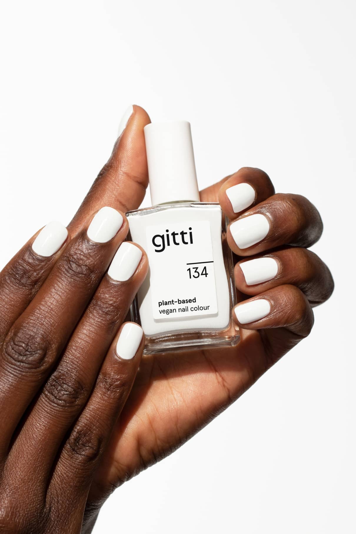 Eine Hand mit weiß lackierten Nägeln hält vor weißem Hintergrund eine Flasche gitti no. 134 vegane Nagelfarbe auf Pflanzenbasis.