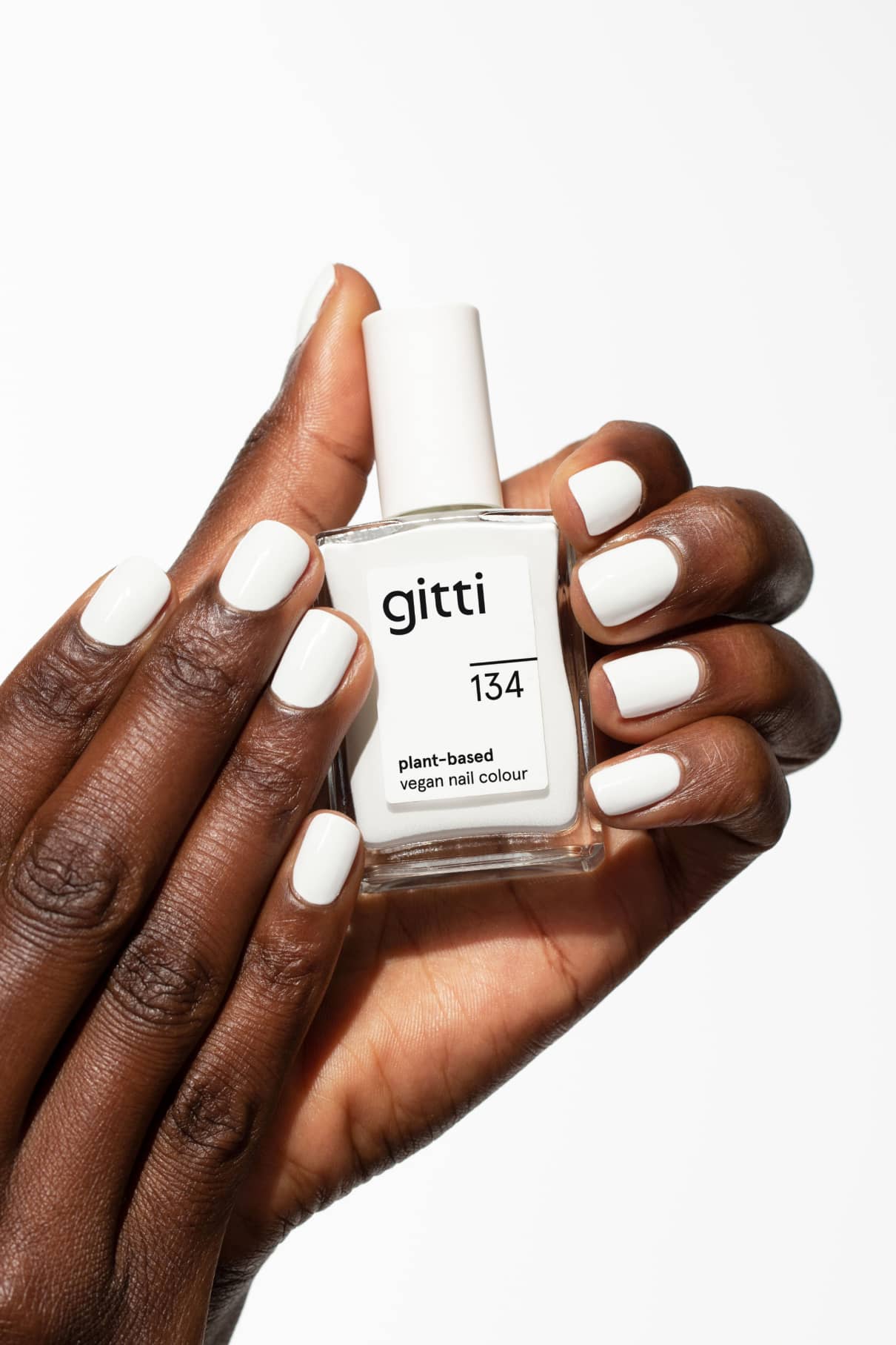 Eine Hand mit weiß lackierten Nägeln hält vor weißem Hintergrund eine Flasche gitti no. 134 vegane Nagelfarbe auf Pflanzenbasis.