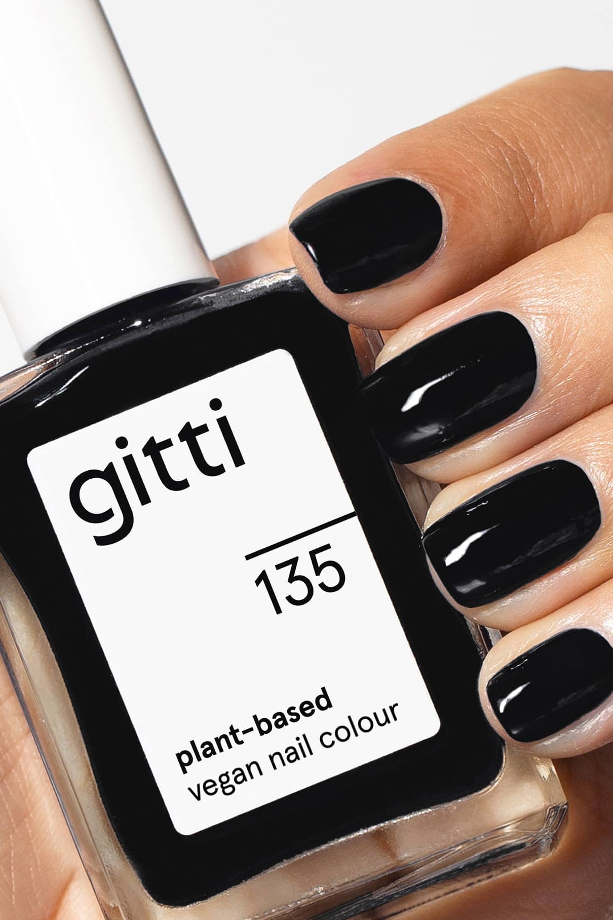 Eine Hand mit kurzen, sauber manikürten und in leuchtendem Schwarz lackierten Nägeln hält ein Fläschchen mit gitti no. 135 veganen Nagellack auf Pflanzenbasis.
