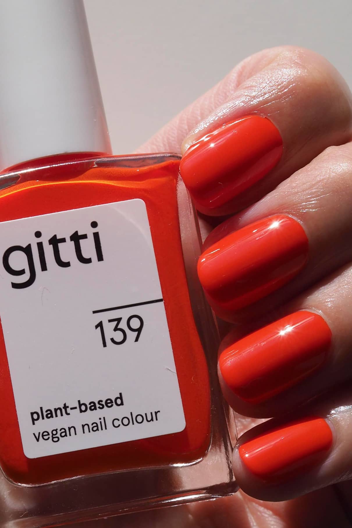 Eine Hand mit sauber manikürten, in leuchtendem Orange-Rot lackierten Nägeln hält eine Flasche gitti no. 139 pflanzlichen, veganen Nagellack in demselben auffälligen Farbton.
