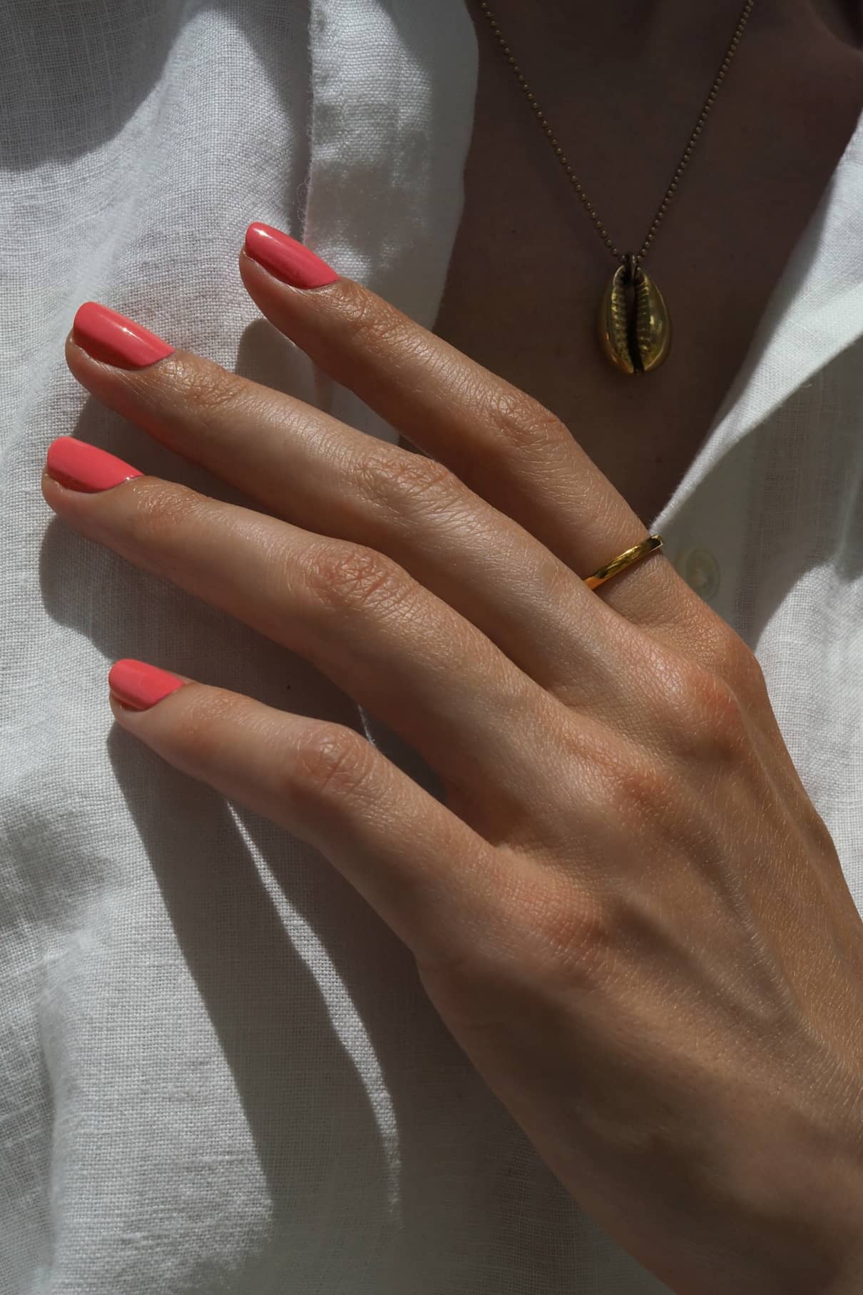Eine Hand mit gitti no. 140 lackierten Nägeln und einem rosigen Unterton trägt einen dünnen Goldring, der auf einem weißen Hemd ruht. Im Hintergrund glänzt dezent eine goldene Halskette mit einem Muschel-Anhänger.