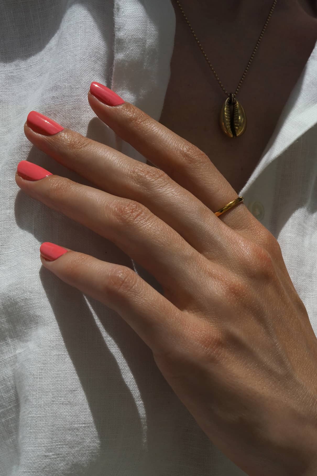 Eine Hand mit gitti no. 140 lackierten Nägeln und einem rosigen Unterton trägt einen dünnen Goldring, der auf einem weißen Hemd ruht. Im Hintergrund glänzt dezent eine goldene Halskette mit einem Muschel-Anhänger.