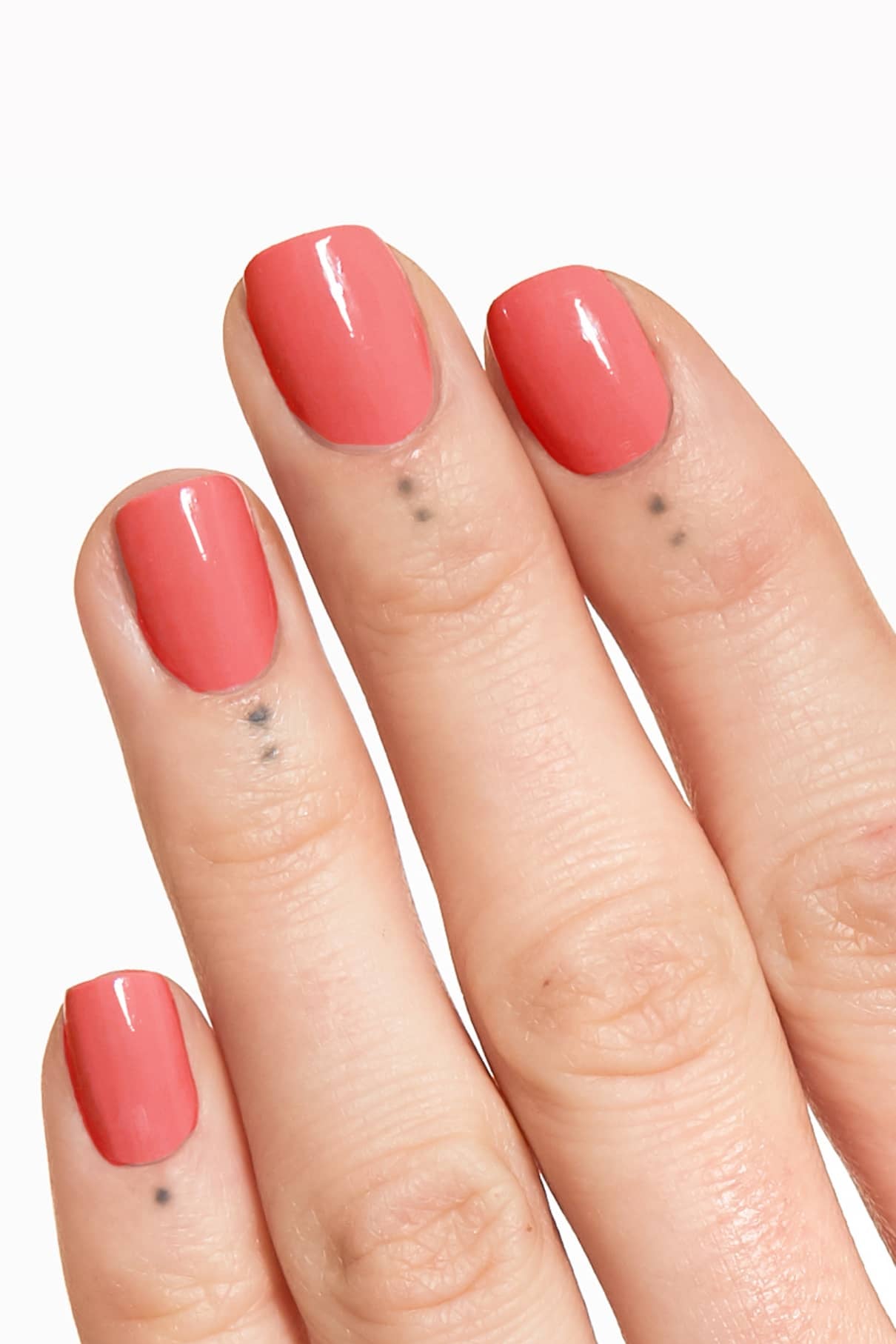 Eine Hand mit kurzen, glänzenden rosa Nägeln in gitti no. 140, mit einem hellen Korallenton mit rosigem Unterton und kleinen schwarzen Punkttattoos unter jedem Fingernagel.