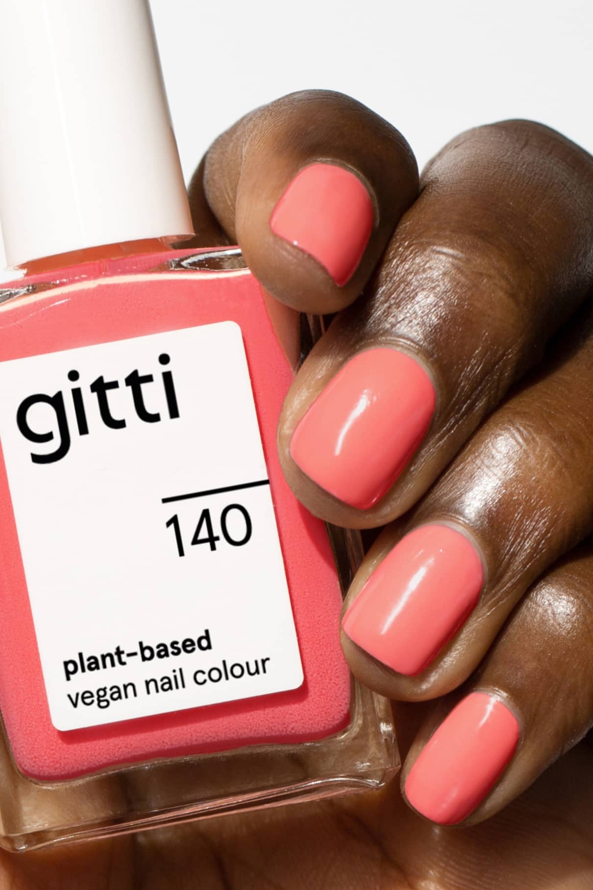 Eine Hand mit korallenrosa lackierten Nägeln hält eine Flasche mit gitti no. 140, eine pflanzliche vegane Nagelfarbe in einem leuchtenden Korallenton mit rosigem Unterton, vor einem schlichten Hintergrund.