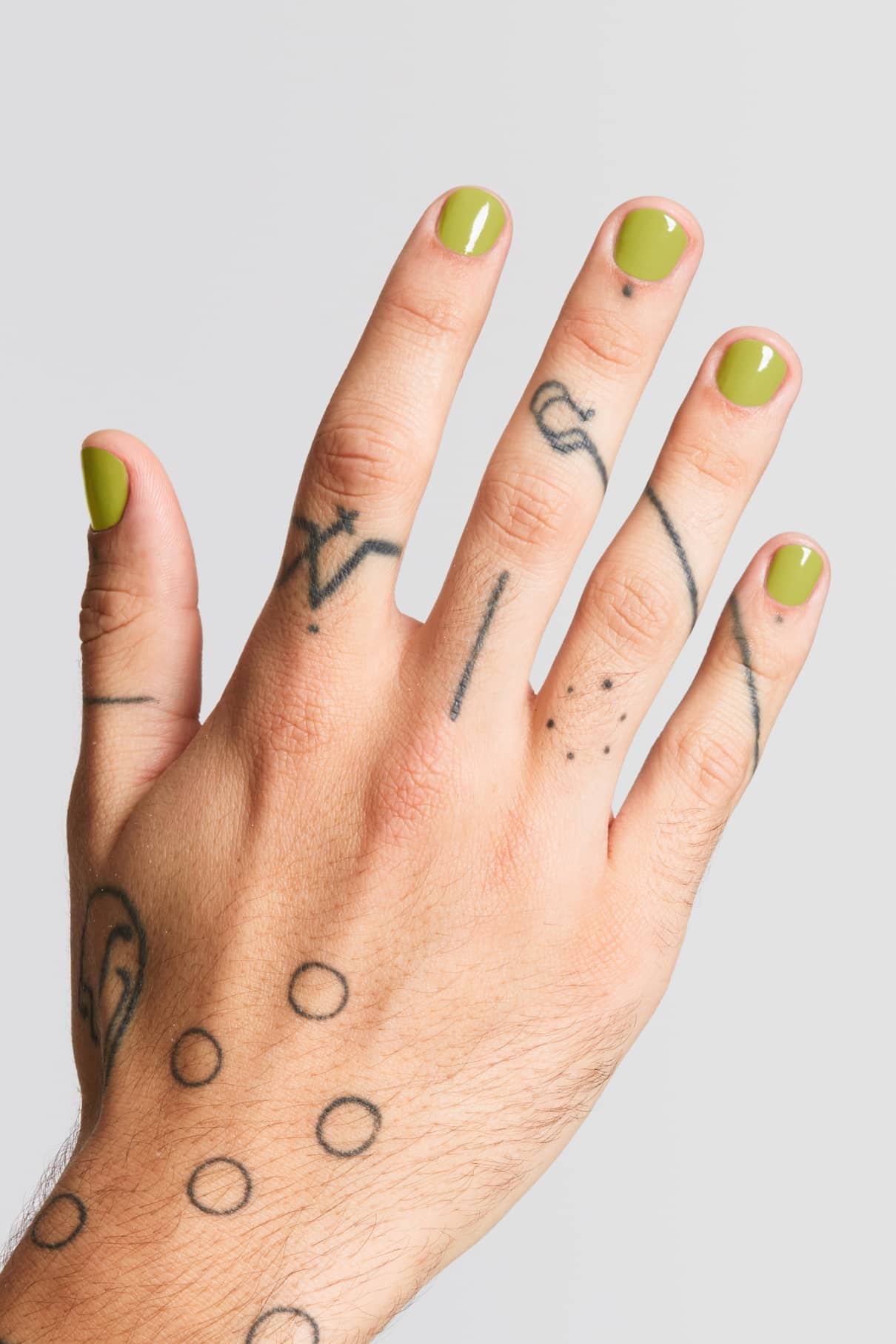 Eine Hand mit Fingernägeln, die in Gitti Nr. 142 (frisches Gelbgrün) und mit schwarzen Strich- und Punkttattoos auf den Fingern und dem Rücken verziert, vor einem schlichten hellen Hintergrund.