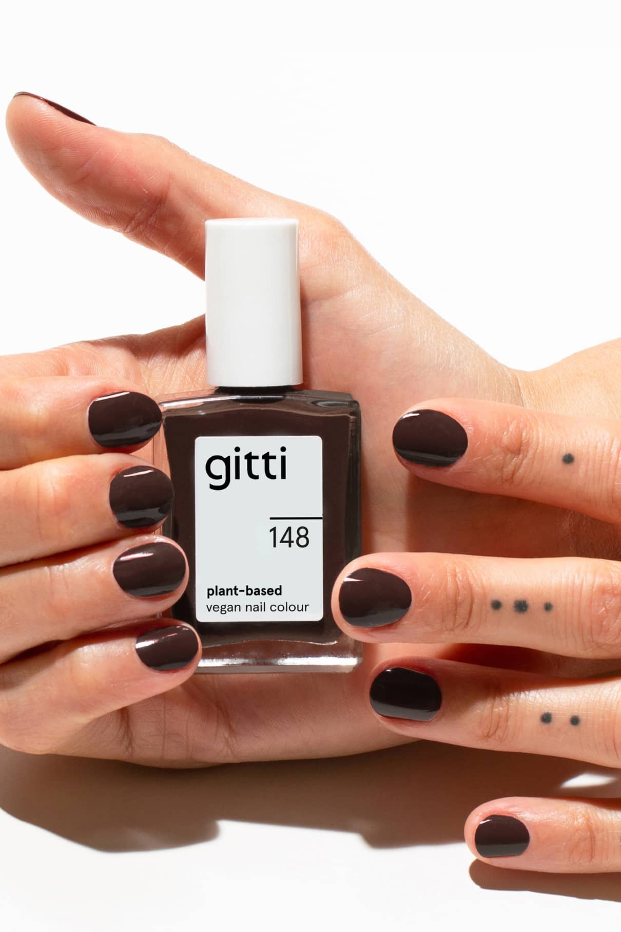 Eine Hand hält eine Flasche mit gitti no. 148, einem veganen Nagellack auf pflanzlicher Basis. Die Nägel sind in einem intensiven Braunton lackiert und auf den Fingern sind kleine Punkttattoos zu sehen.