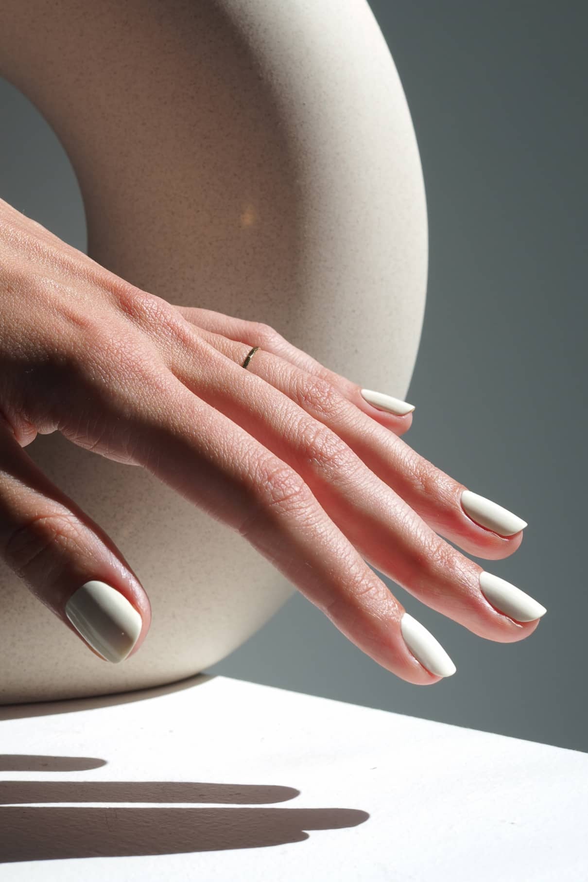 Eine Hand mit kurzen, sauber manikürten Nägeln, die in gitti no. 157, einem cremefarbenen Farbton, ruht auf einer Fläche mit einem einfachen Ring; im Hintergrund setzt ein cremefarbenes, gebogenes Keramikobjekt einen subtilen Akzent.