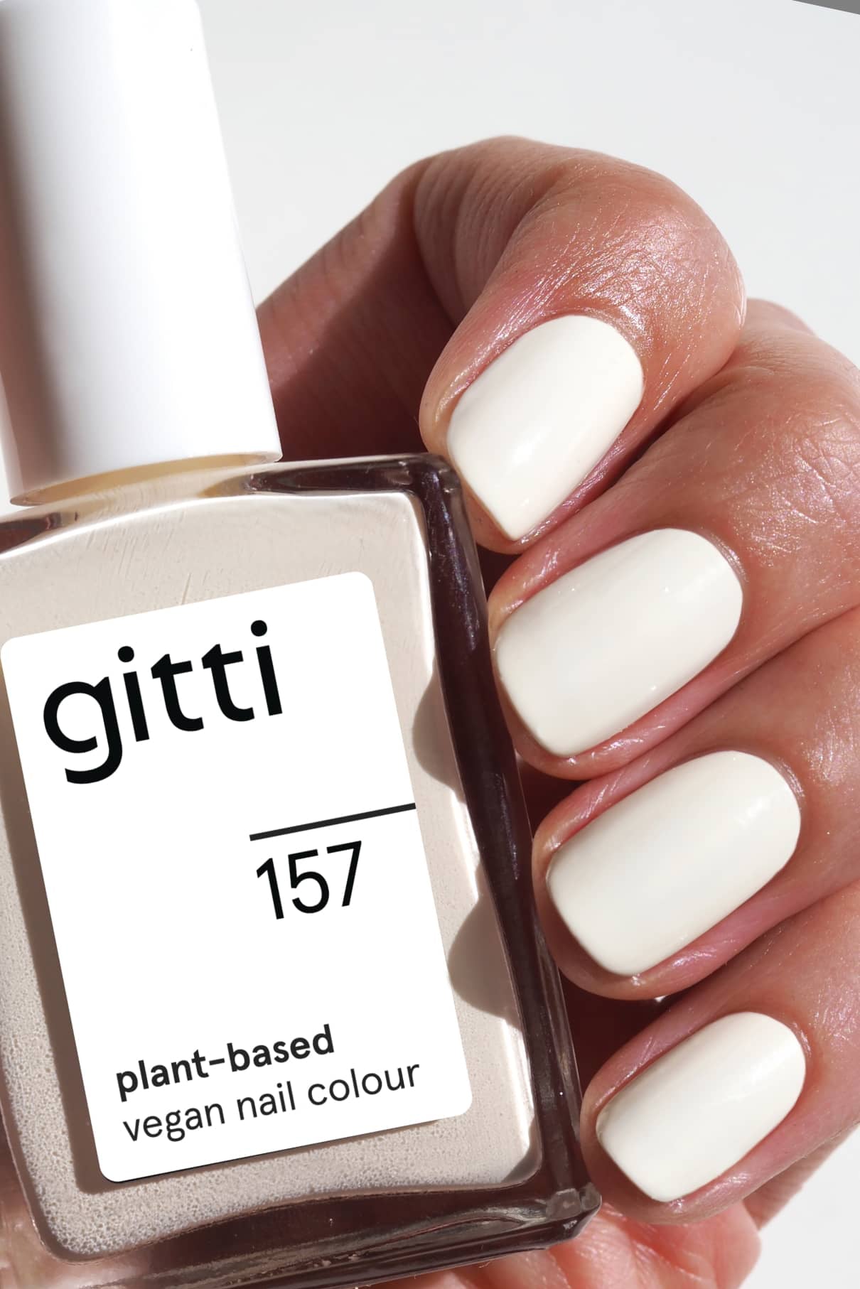 Eine Hand mit kurzen, sauber manikürten Nägeln, die in glänzendem Off-White-Lack lackiert sind, hält eine Flasche gitti no. 157 pflanzlicher veganer Nagellack.