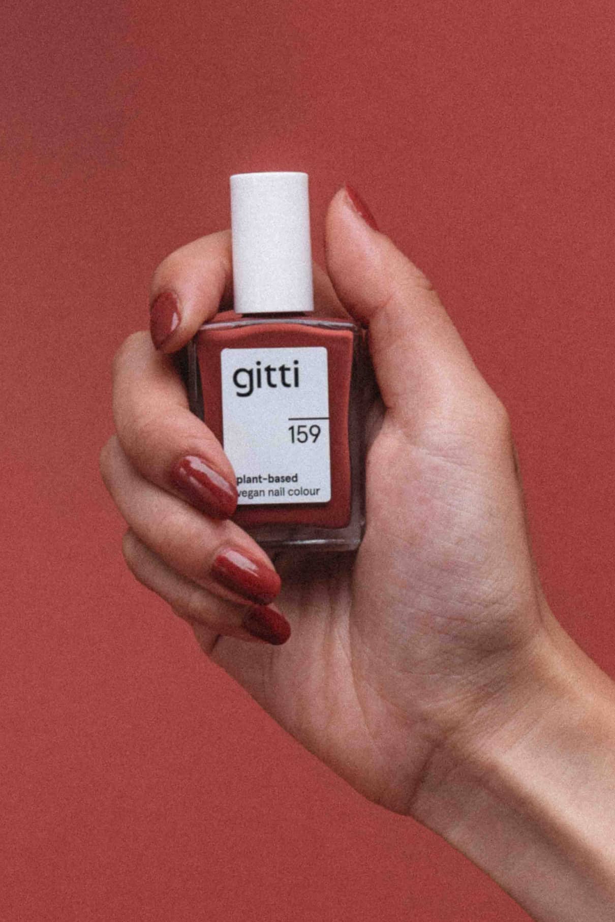 Eine Hand mit rot lackierten Nägeln in pudrigem Rot hält ein Fläschchen mit gitti no. 159 pflanzlicher veganer Nagelfarbe vor einem passenden roten Hintergrund.