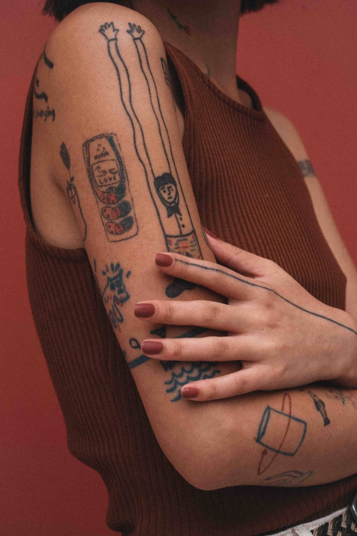 Eine Person mit kurzen dunklen Haaren und einem ärmellosen braunen Oberteil zeigt einen Arm, der mit lebhaften Tattoos bedeckt ist - lange Arme, eine Dose, ein Gesicht, abstrakte Motive - mit einem Hauch von pudrigem Rot, inspiriert von gitti no. 159 in den lebhaften Motiven.