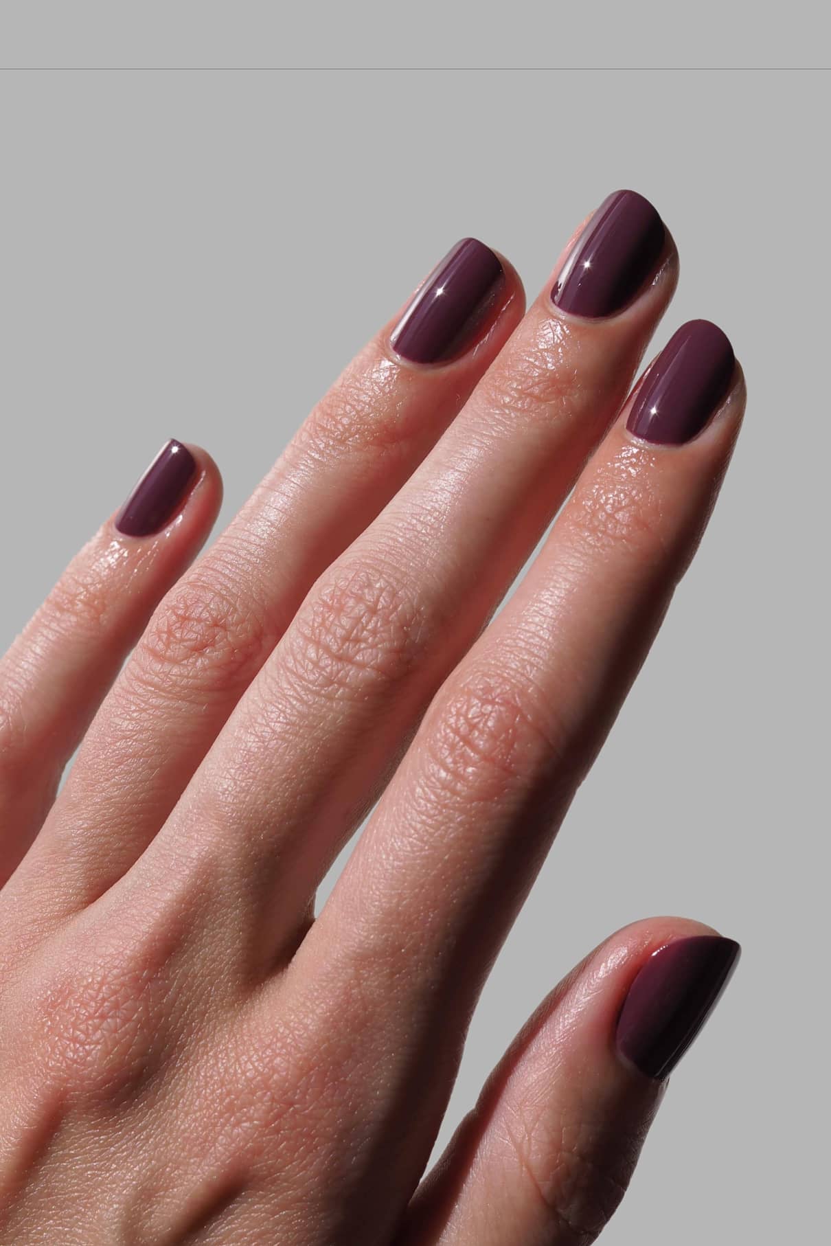 Eine Hand mit kurzen, sauber manikürten Nägeln, lackiert in glänzendem gitti no. 164, einem rötlichen Lila-Lack mit blauen Untertönen, dargestellt vor einem schlichten grauen Hintergrund.