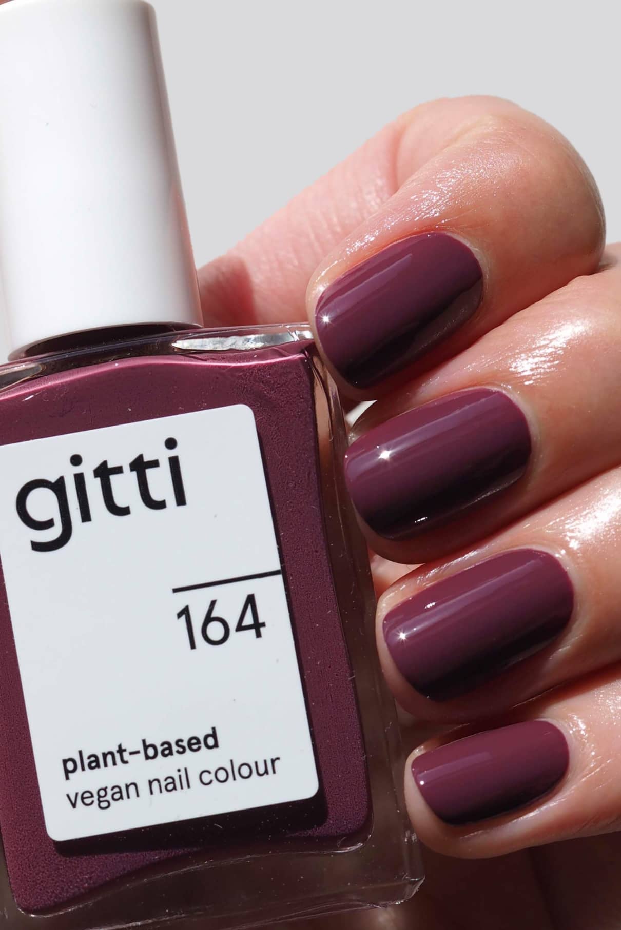 Eine Hand mit kurzen, sauber manikürten Nägeln, die in einem rötlich-violetten Mauveton lackiert sind, hält eine Flasche gitti no. 164 pflanzlichen veganen Nagellack.