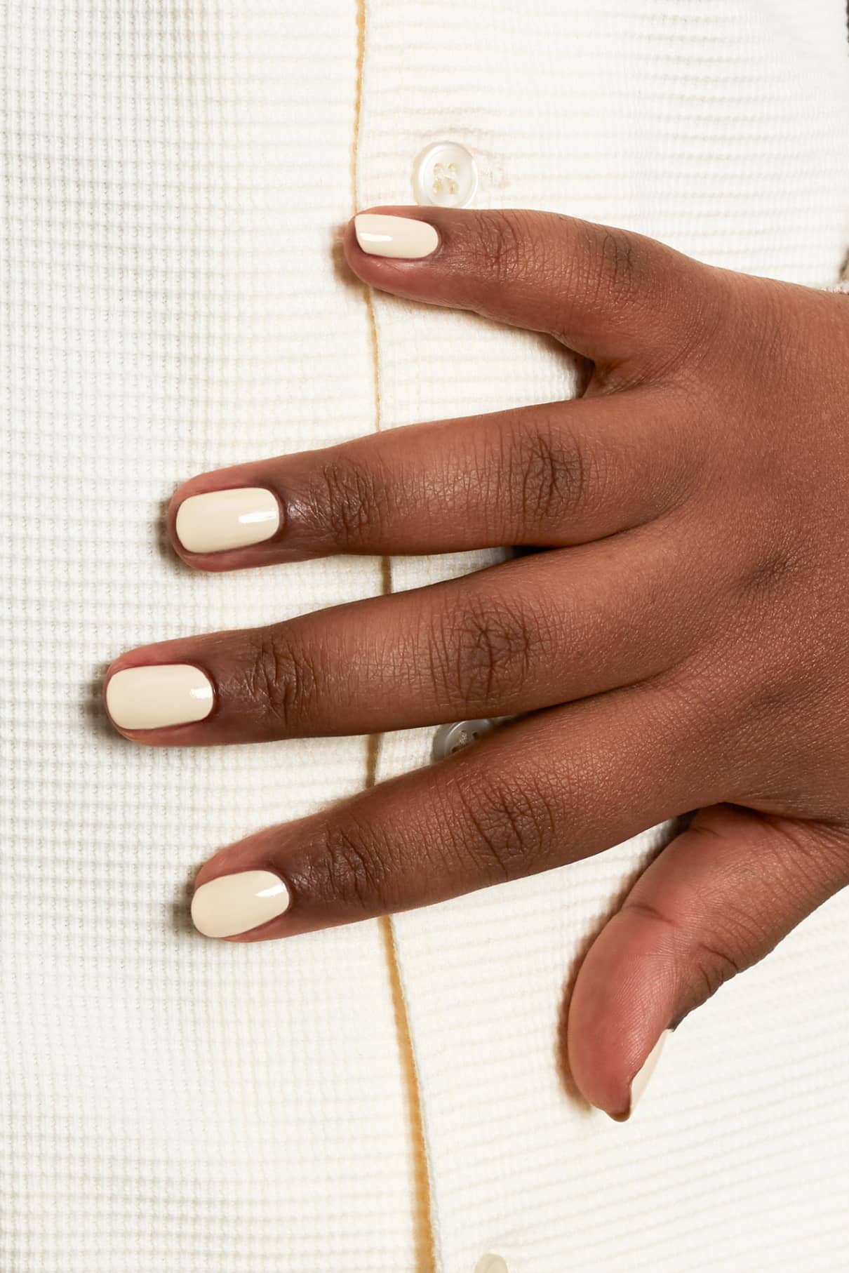 Eine Hand mit kurzen, sauber manikürten Nägeln, lackiert in gitti no. 165, einem hellen Cremeton mit gelben Untertönen, ruht auf einem strukturierten weißen Stoff mit Knöpfen.