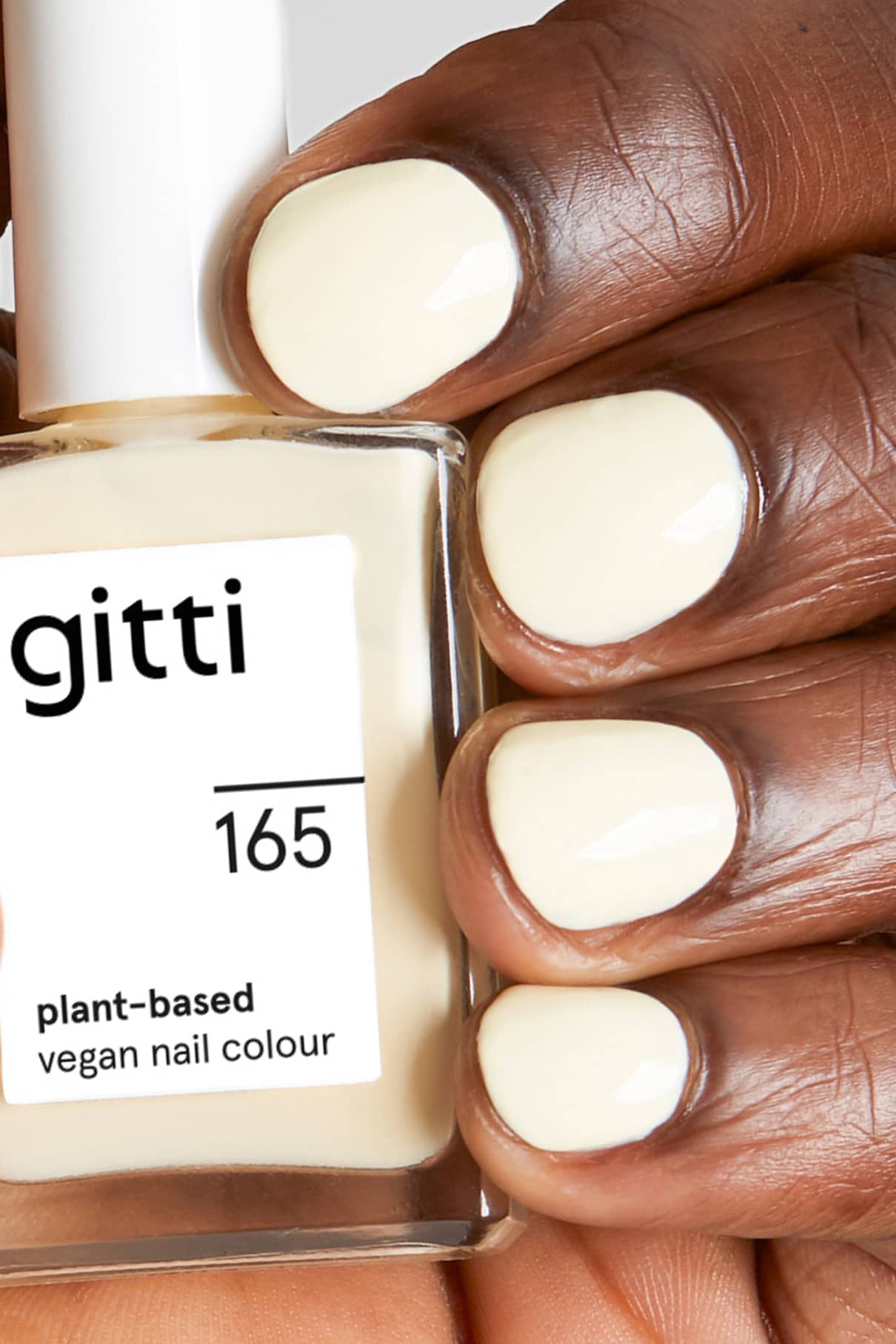 Eine Hand mit kurzen, sauber manikürten Nägeln, die in einem hellen Cremeton mit gelben Untertönen lackiert sind, hält eine Flasche gitti no. 165 veganen Nagellack auf Pflanzenbasis.