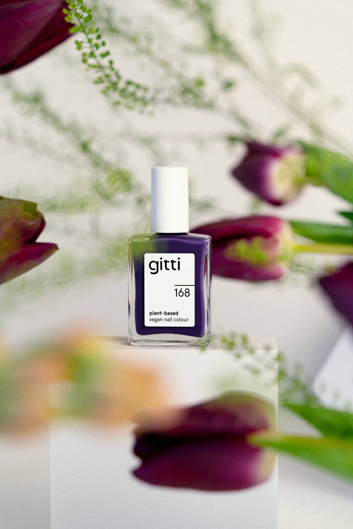 Eine Flasche gitti no. 168 pflanzliche vegane Nagelfarbe in dunklem Lila ruht auf einem weißen Block, um den herum verschwommene lila Blüten und grüne Stängel zu sehen sind.
