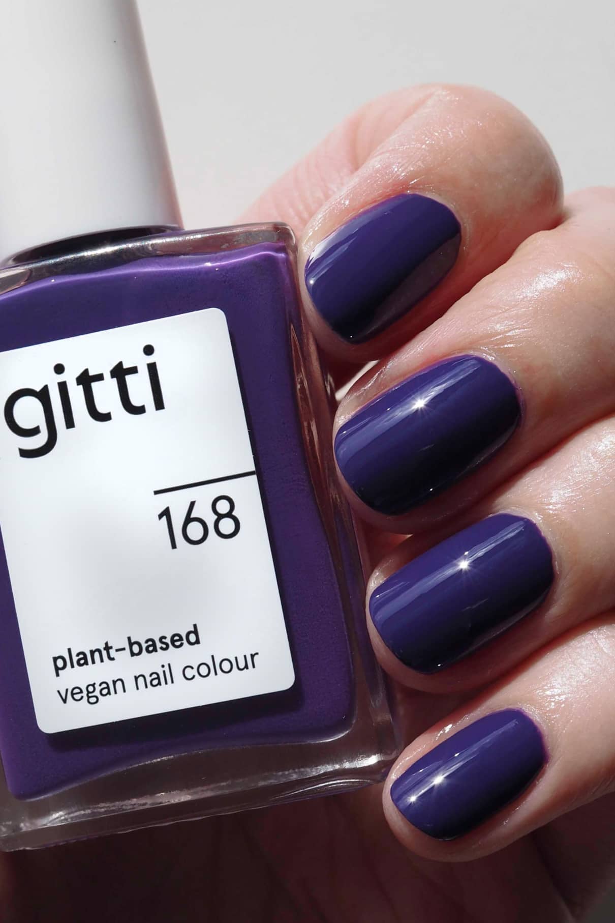 Eine Hand mit kurzen, sauber manikürten Nägeln, die glänzend dunkellila lackiert sind, hält eine Flasche gitti no. 168, einem veganen Nagellack auf pflanzlicher Basis in demselben satten Lila-Ton.