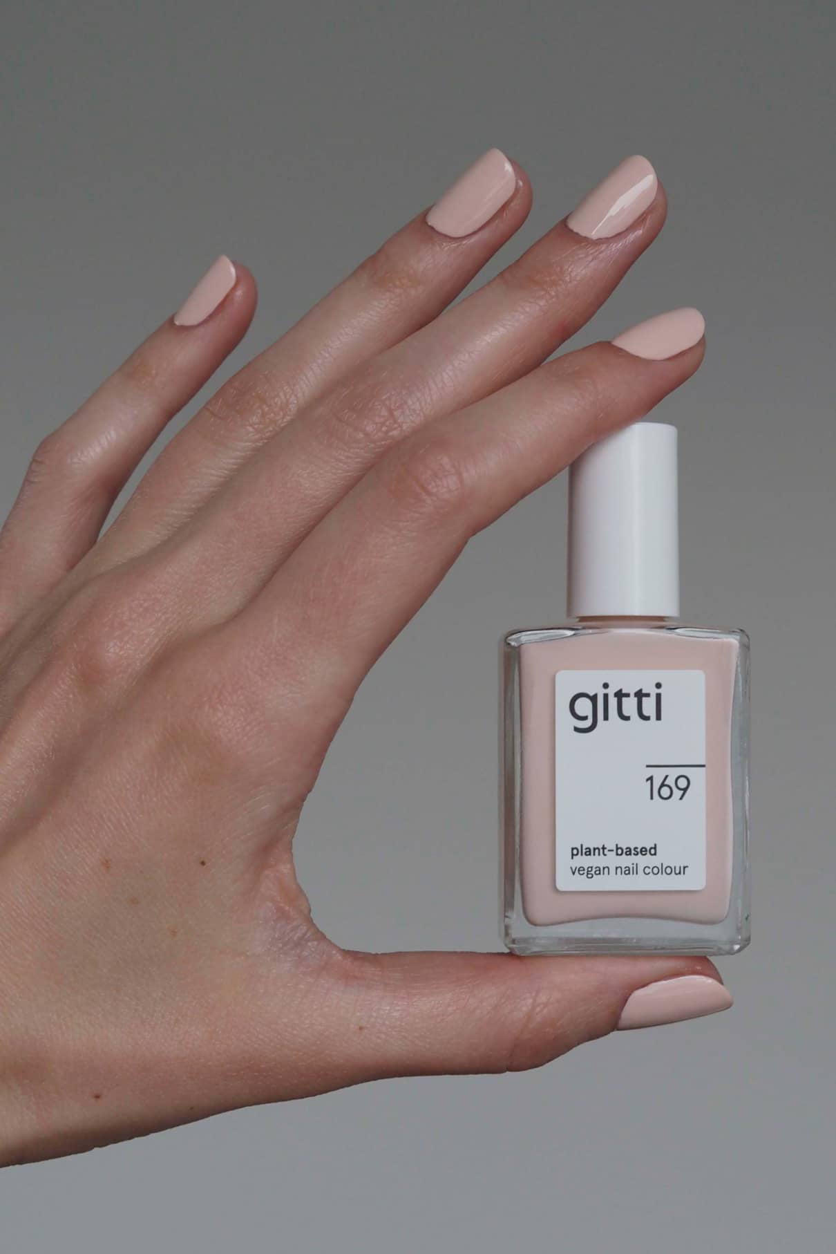 Eine Hand mit sauber manikürten, in zartem Roséton-Rosa lackierten Nägeln hält eine Flasche gitti no. 169 pflanzlicher veganer Nagellack vor einem schlichten Hintergrund.