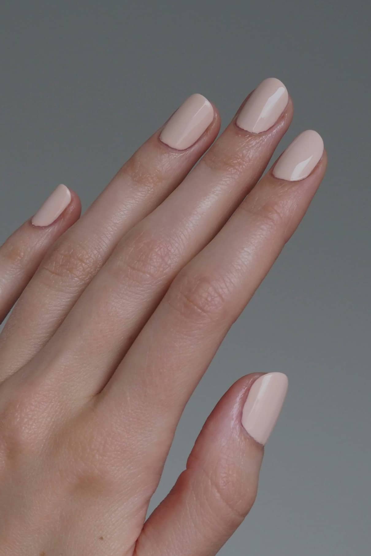 Eine Hand mit sauber manikürten Nägeln, lackiert in gitti no. 169, einem glänzenden, von Roséton inspirierten, blassen Nude-Ton, vor einem schlichten grauen Hintergrund.