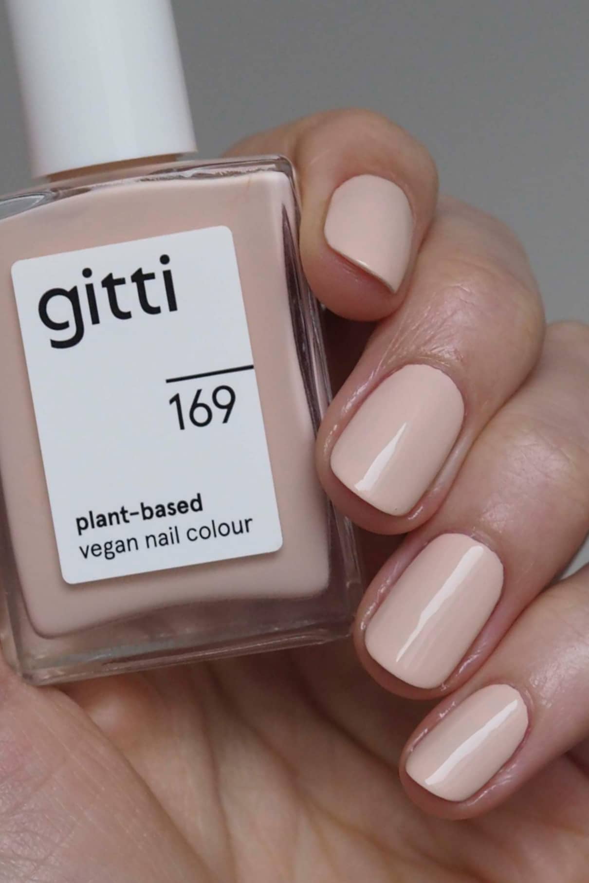 Eine Hand mit kurzen, sauber manikürten Nägeln, lackiert in einem blassen Nude-Ton von gitti no. 169 lackiert sind, hält eine Flasche dieses veganen Nagellacks auf Pflanzenbasis.