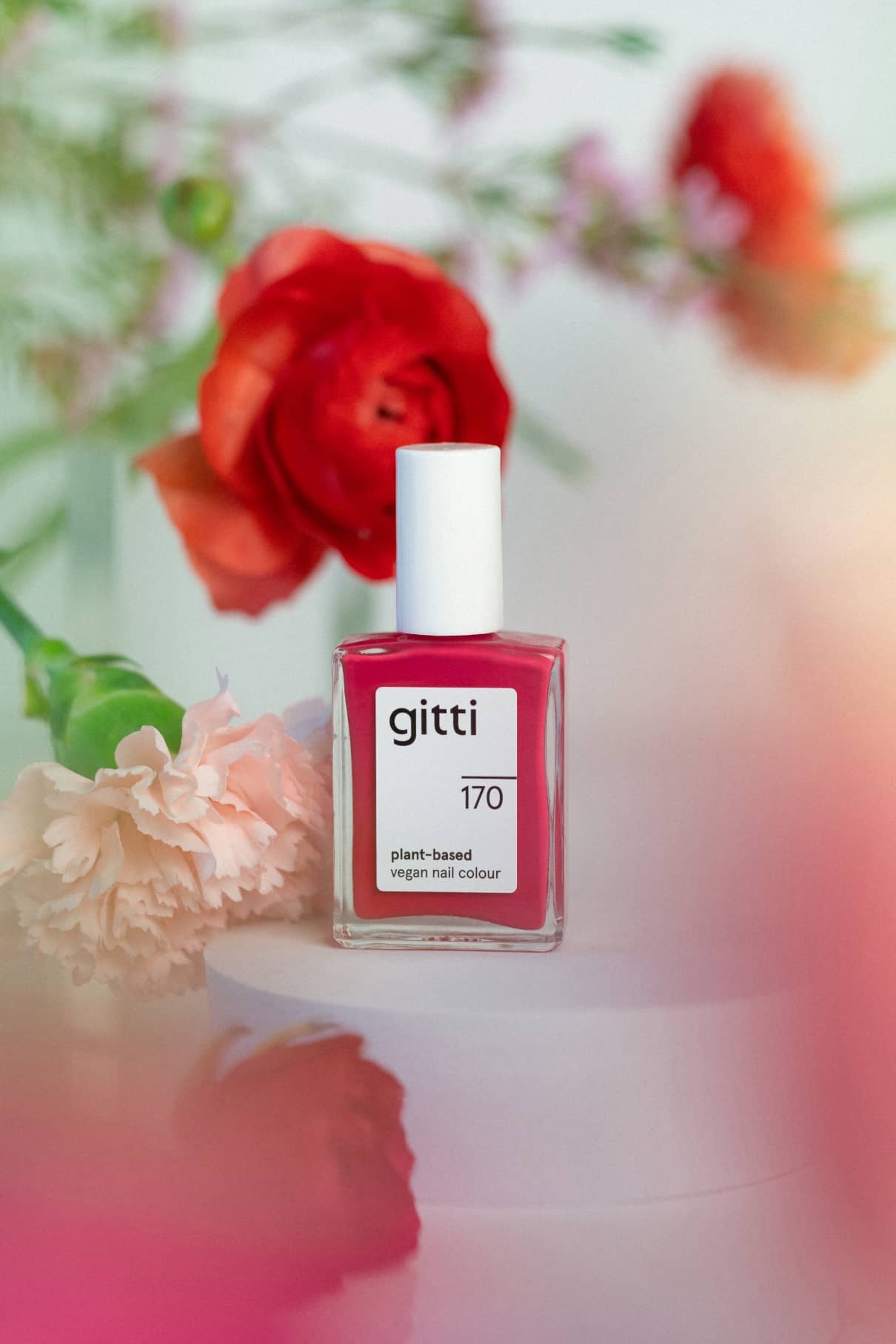 Eine Flasche gitti no. 170 vegane Nagelfarbe auf Pflanzenbasis in Magenta-Rot steht auf einem weißen Podest mit roten und rosa Blumen im Hintergrund.