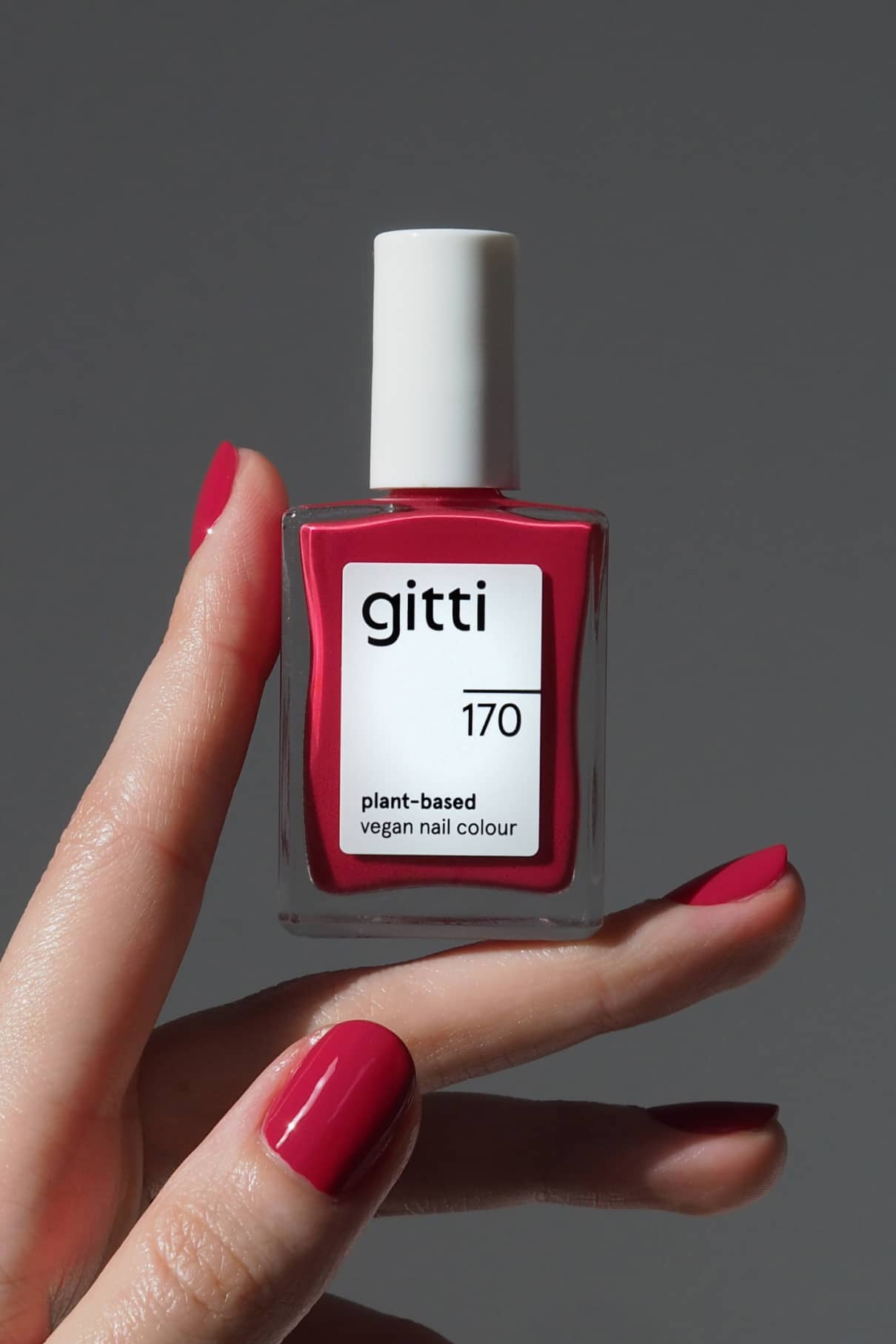 Eine Hand mit rot lackierten Nägeln hält eine Flasche von gitti no. 170, einem veganen Nagellack auf Pflanzenbasis in Magenta-Rot, vor einem schlichten grauen Hintergrund.