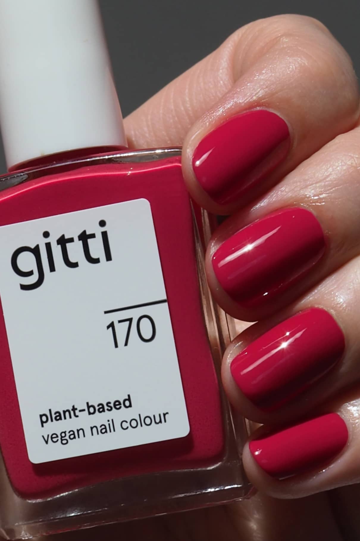 Eine Hand mit sauber manikürten Nägeln in einem glänzenden Magenta-Rot-Ton hält eine Flasche gitti no. 170 veganen Nagellack auf Pflanzenbasis.