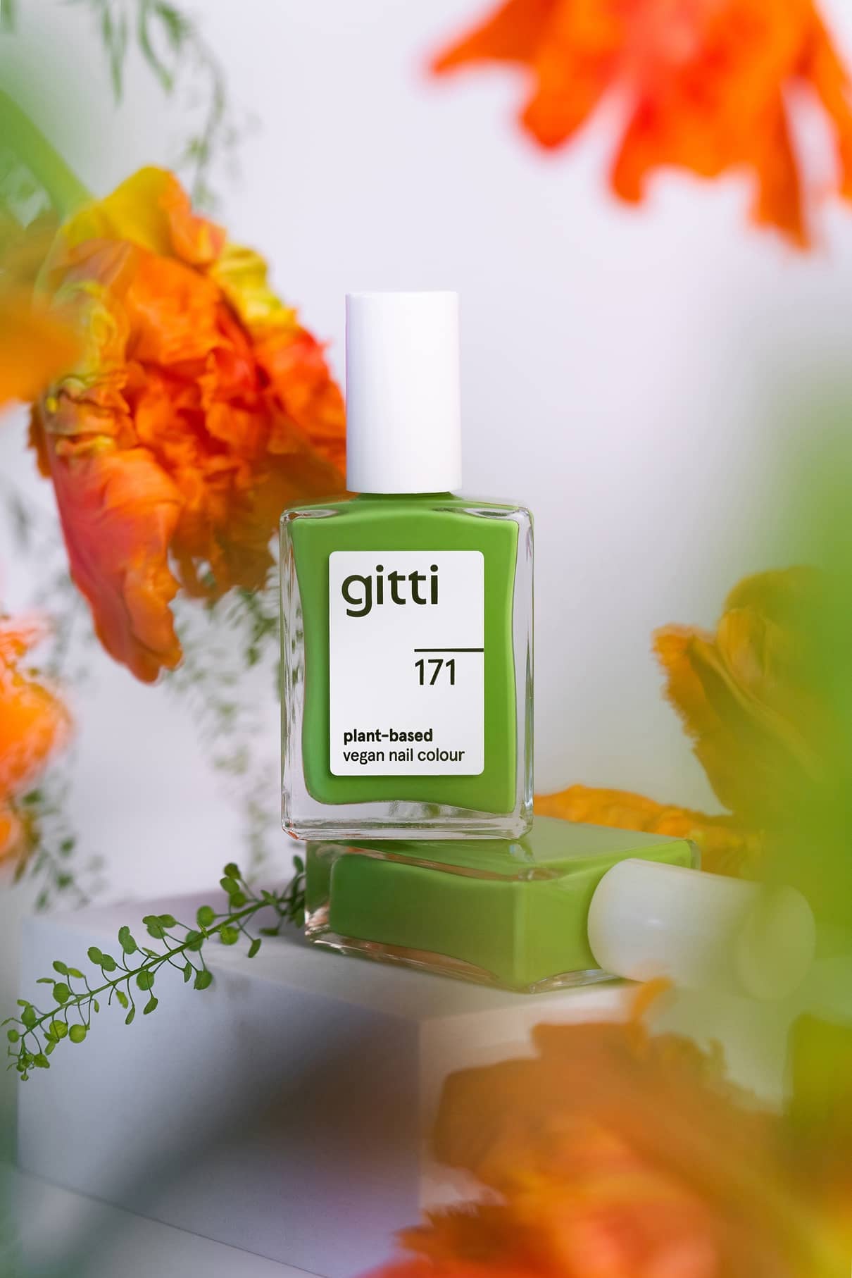Eine Flasche von gitti no. 171, eine pflanzliche, vegane Nagelfarbe in frischem Grasgrün, ist zwischen orangefarbenen Blumen und üppigem Grün zu sehen.