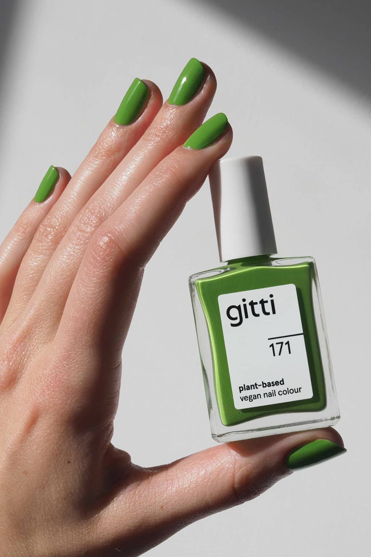 Eine Hand mit grün lackierten Nägeln hält eine Flasche gitti no. 171 pflanzlicher, veganer Nagelfarbe, die perfekt zu ihrem leuchtenden Farbton frisches Grasgrün passt.