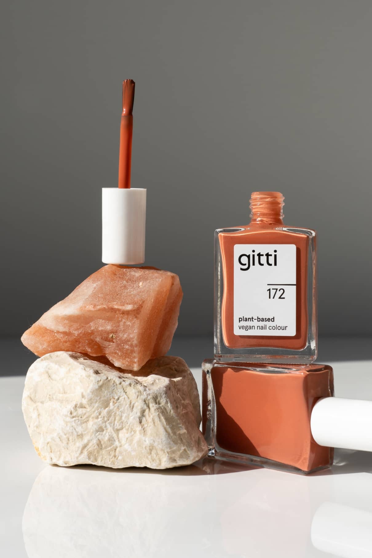 Eine Flasche von gitti no. 172 pflanzlicher, veganer Nagellack im Farbton Terracottarot mit sanften, warmen Untertönen wird geöffnet und auf zwei Steinen balanciert, während eine andere Flasche auf der Seite liegt.