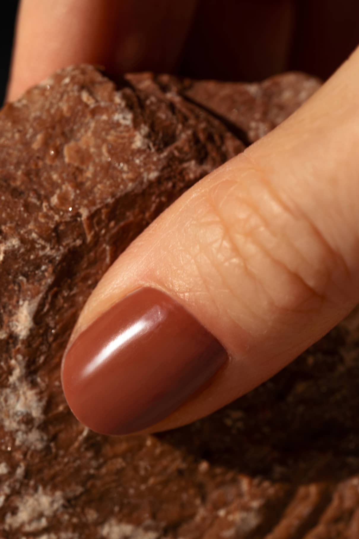Nahaufnahme eines Daumens mit gitti no. 172, einem sanften terrakottabraunen Nagellack mit warmen Untertönen, der sanft auf einen strukturierten braunen Stein gedrückt wird.