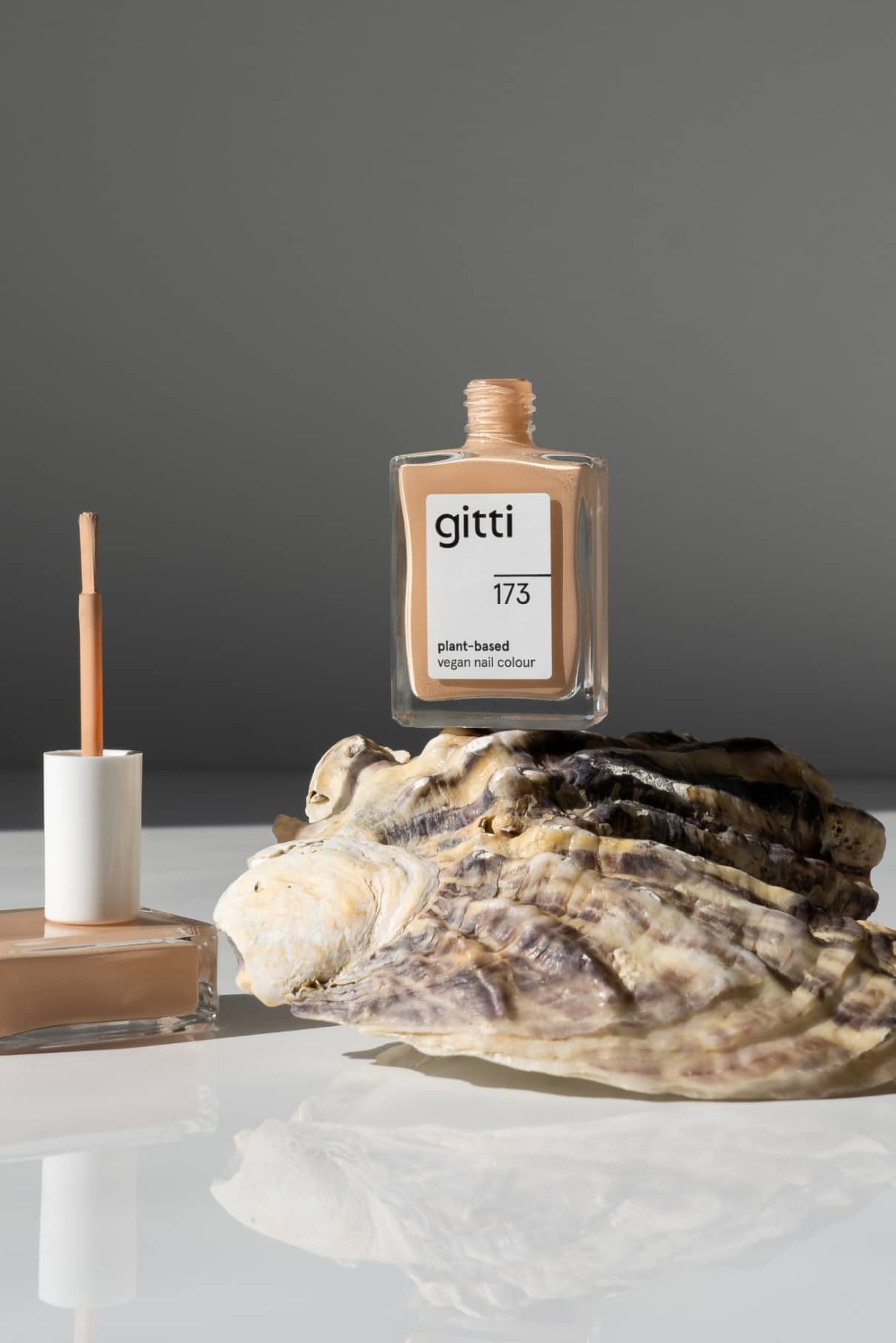 Eine Flasche gitti no. 173 pflanzliche vegane Nagelfarbe in hellem Apricot mit warmen Untertönen steht auf einer großen Muschel, neben einer offenen Nagellackflasche und einem Pinsel, vor einem schlichten grauen Hintergrund.
