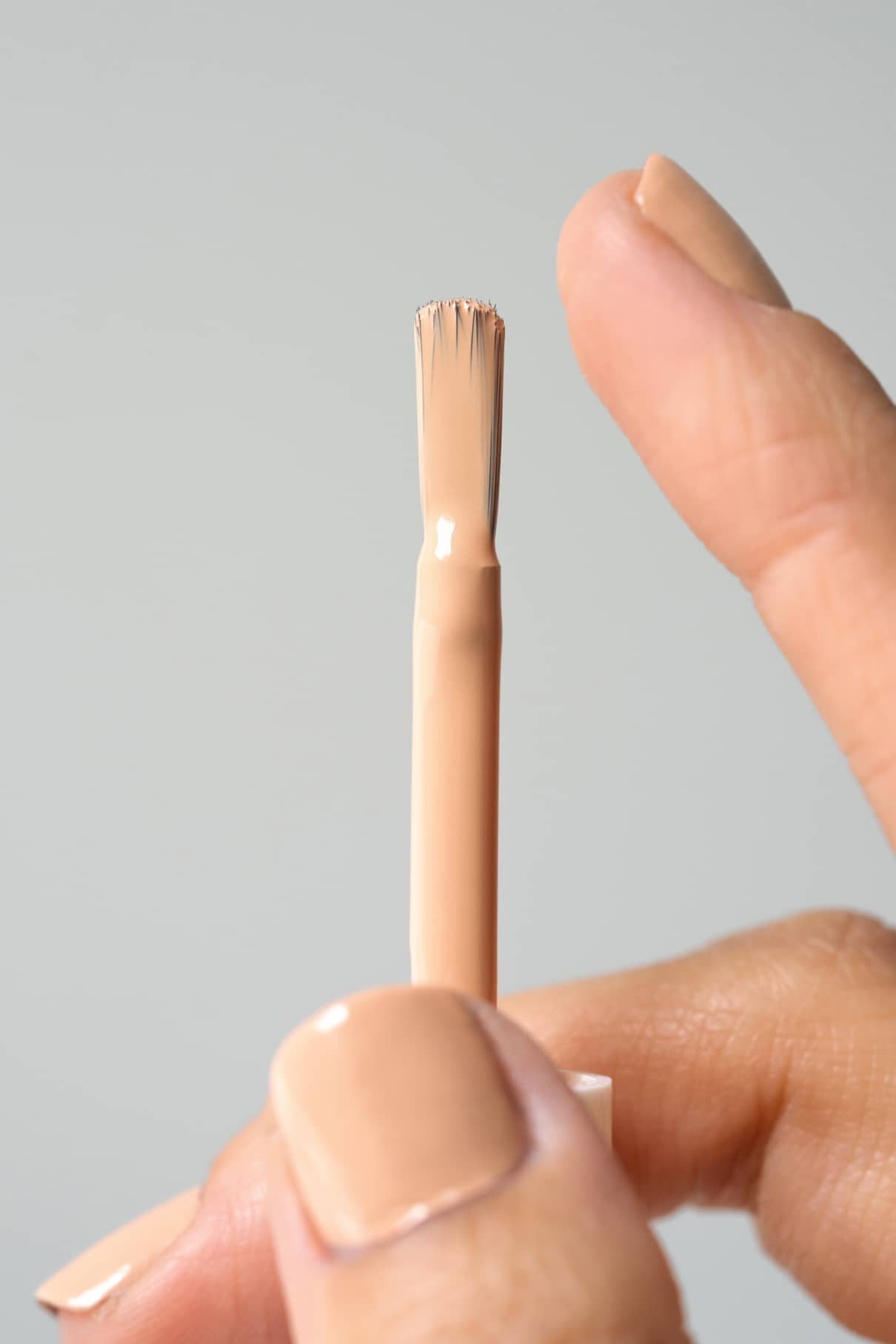 Eine Hand hält einen gitti no. 173 Nagellackpinsel mit weichem Apricot-Farbton neben einem nude lackierten Fingernagel mit warmen Untertönen, vor einem schlichten Hintergrund.