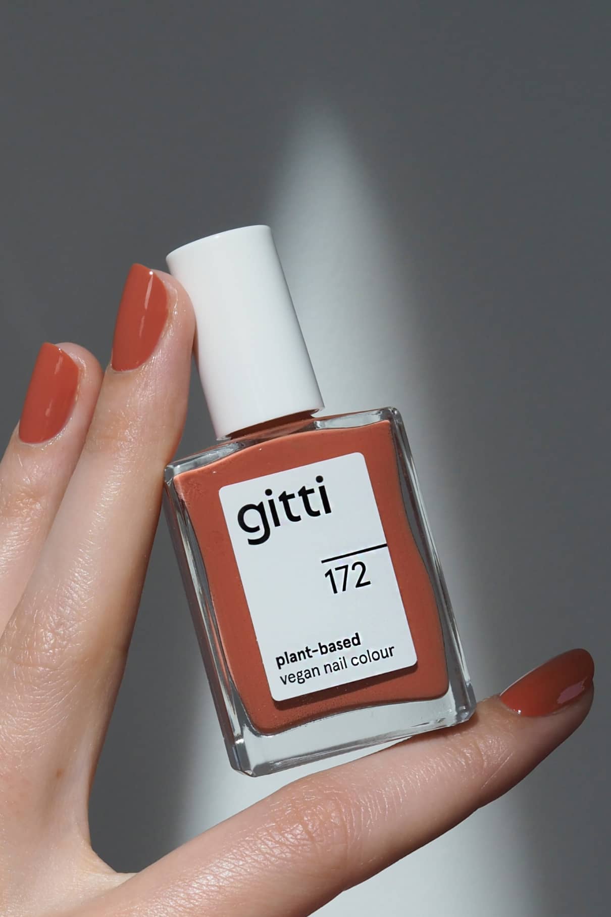 Eine Hand mit manikürten Nägeln hält eine Flasche von gitti no. 172, einem pflanzlichen, veganen Nagellack in dem sanften, rotbraunen Farbton Terracottarot mit warmen Untertönen.
