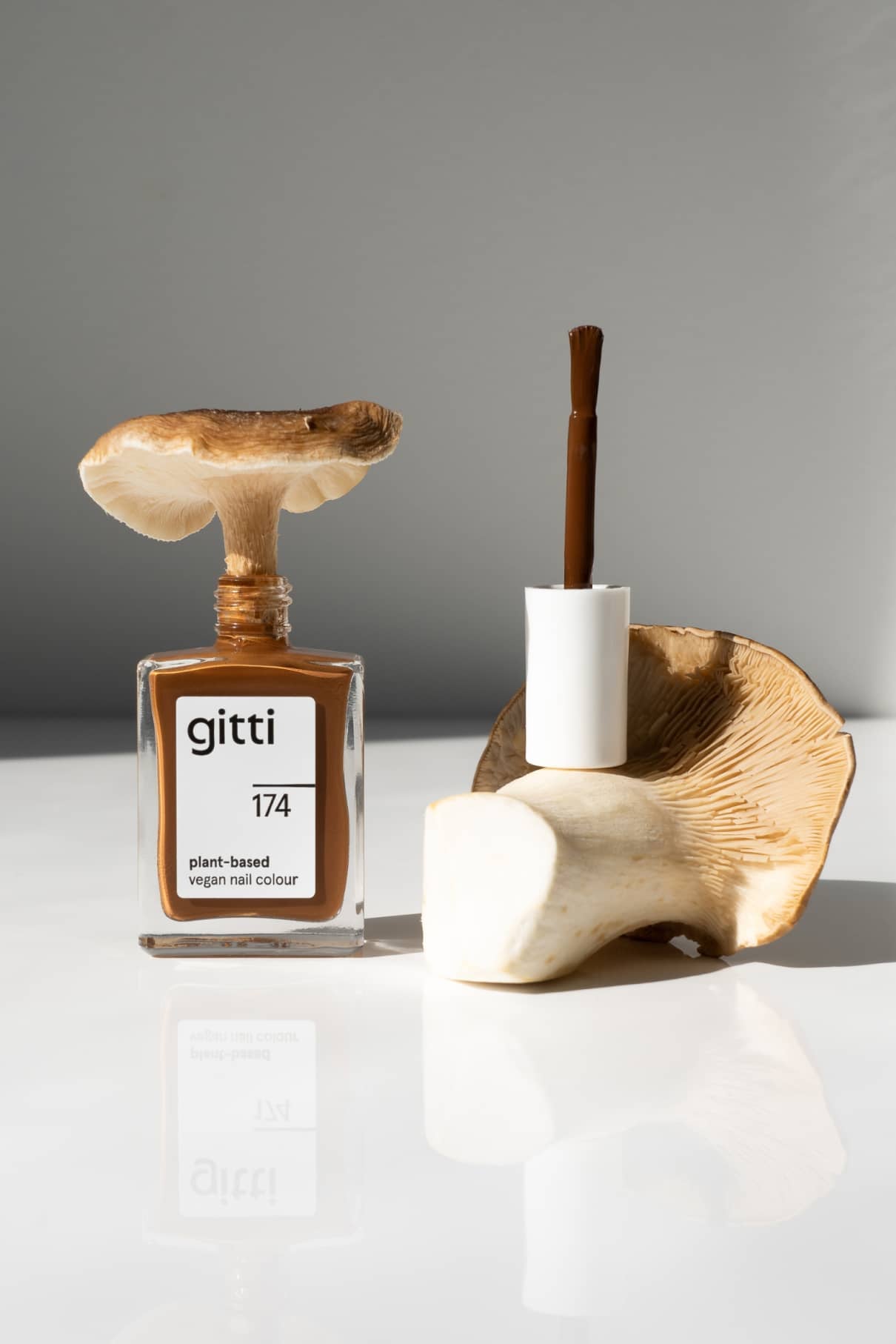 Eine Flasche mit gitti no. 174 pflanzlicher veganer Nagellack (ein erdiges Braun mit gelben Untertönen) steht auf einer weißen Fläche, die von einem Pilz gekrönt wird, während ein weiterer Pilz den Pinsel hält.