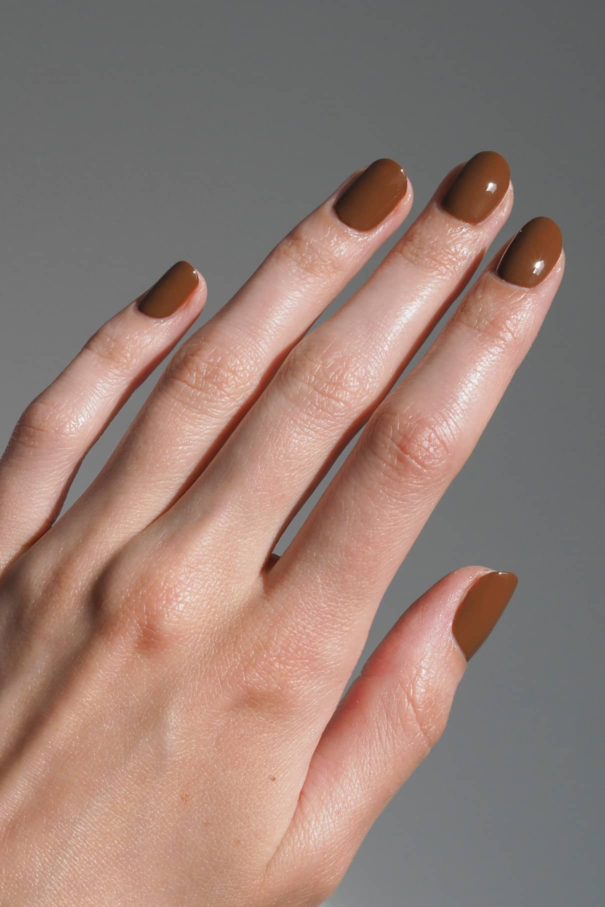 Eine Hand mit kurzen, mandelförmigen Nägeln, lackiert in glänzendem Gitti Nr. 174, einem erdigen Braunton, vor einem neutralen Hintergrund.