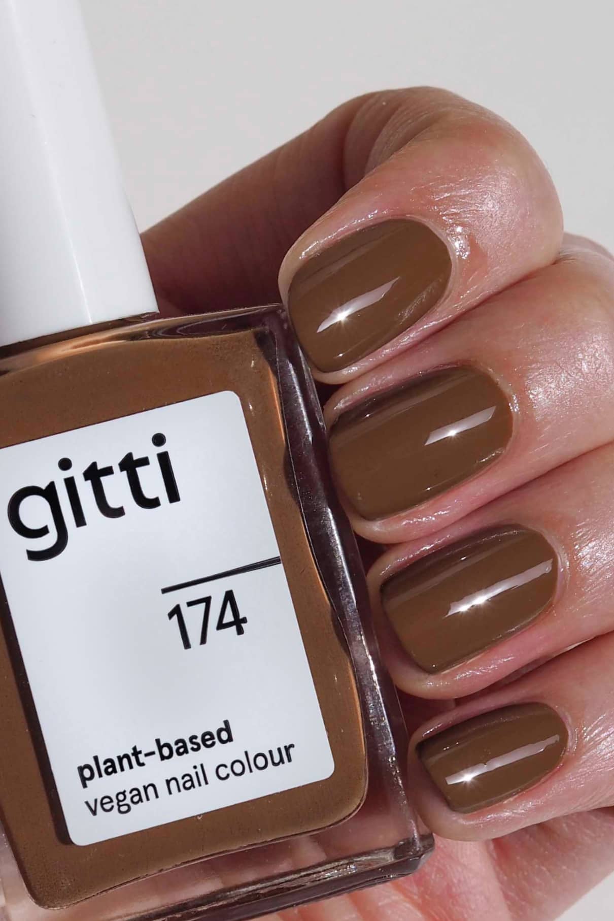 Eine Hand mit kurzen, sauber manikürten Nägeln, die in einem erdigen Braunton lackiert sind, hält ein Fläschchen mit gitti no. 174 pflanzlichen veganen Nagellack, der perfekt auf die Nagelfarbe abgestimmt ist.