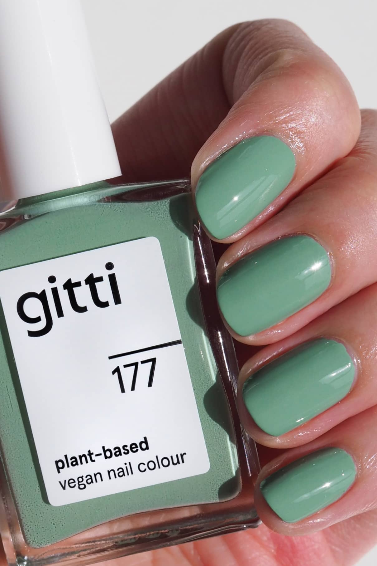Eine Hand mit kurzen, in einem sanften Grünton lackierten Nägeln hält eine Flasche gitti no. 177 veganen Nagellack auf Pflanzenbasis.