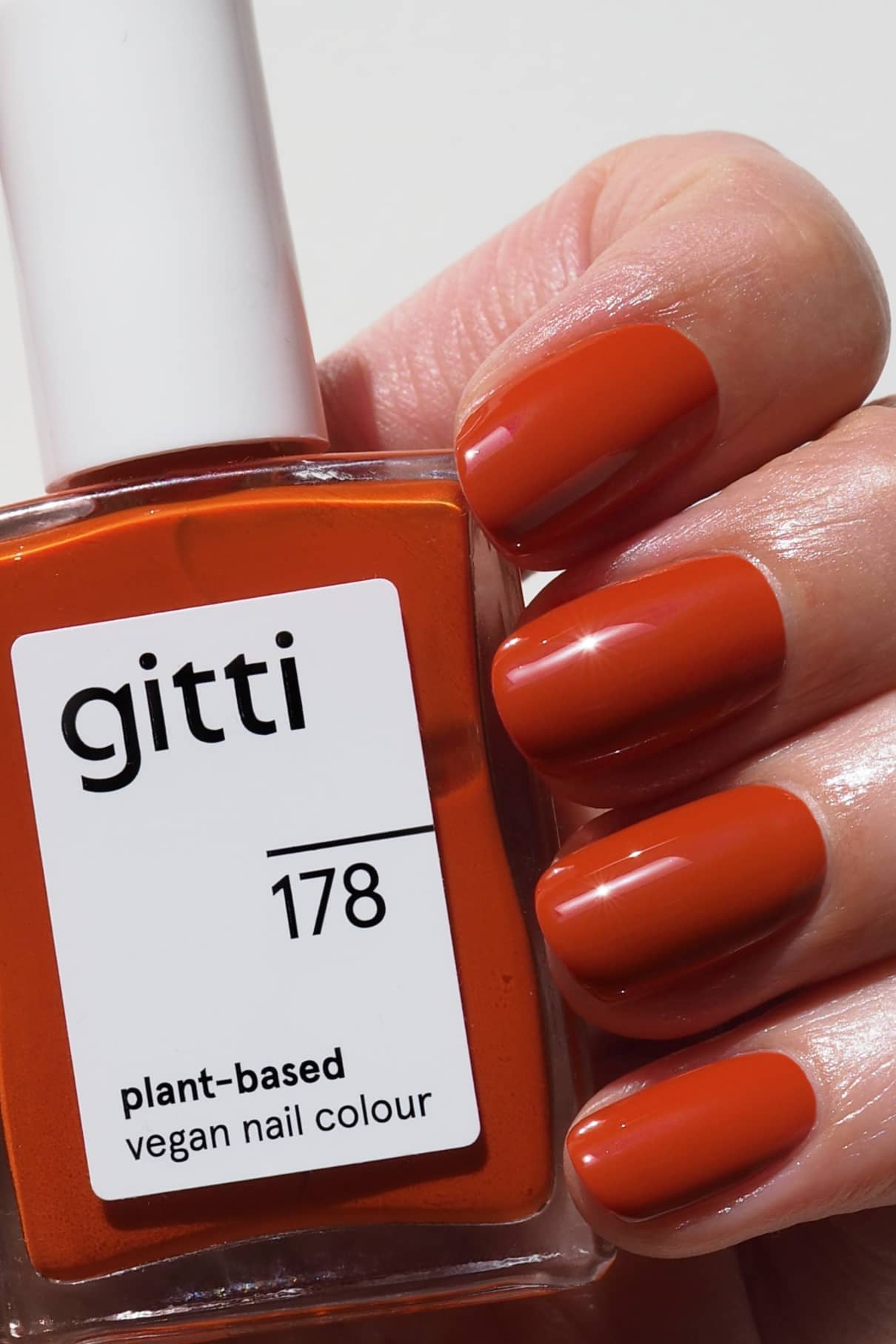 Eine Hand mit sauber manikürten Nägeln in einem warmen, gesättigten Orangerot-Ton hält eine Flasche gitti no. 178 veganen Nagellack auf Pflanzenbasis.