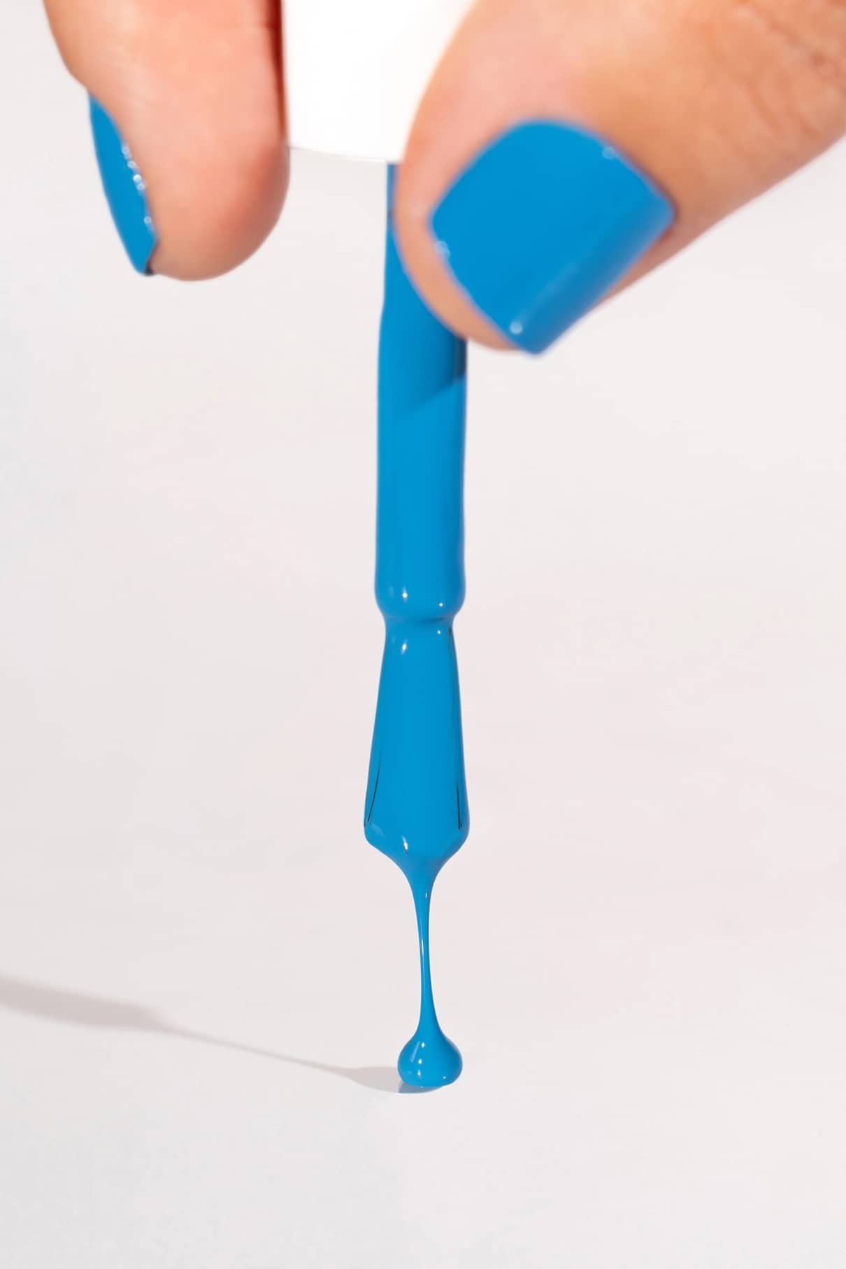Eine Hand mit gitti no. 179 Nagellack hält einen Pinsel, blauer Lack tropft von den Borsten vor einem schlichten Hintergrund.