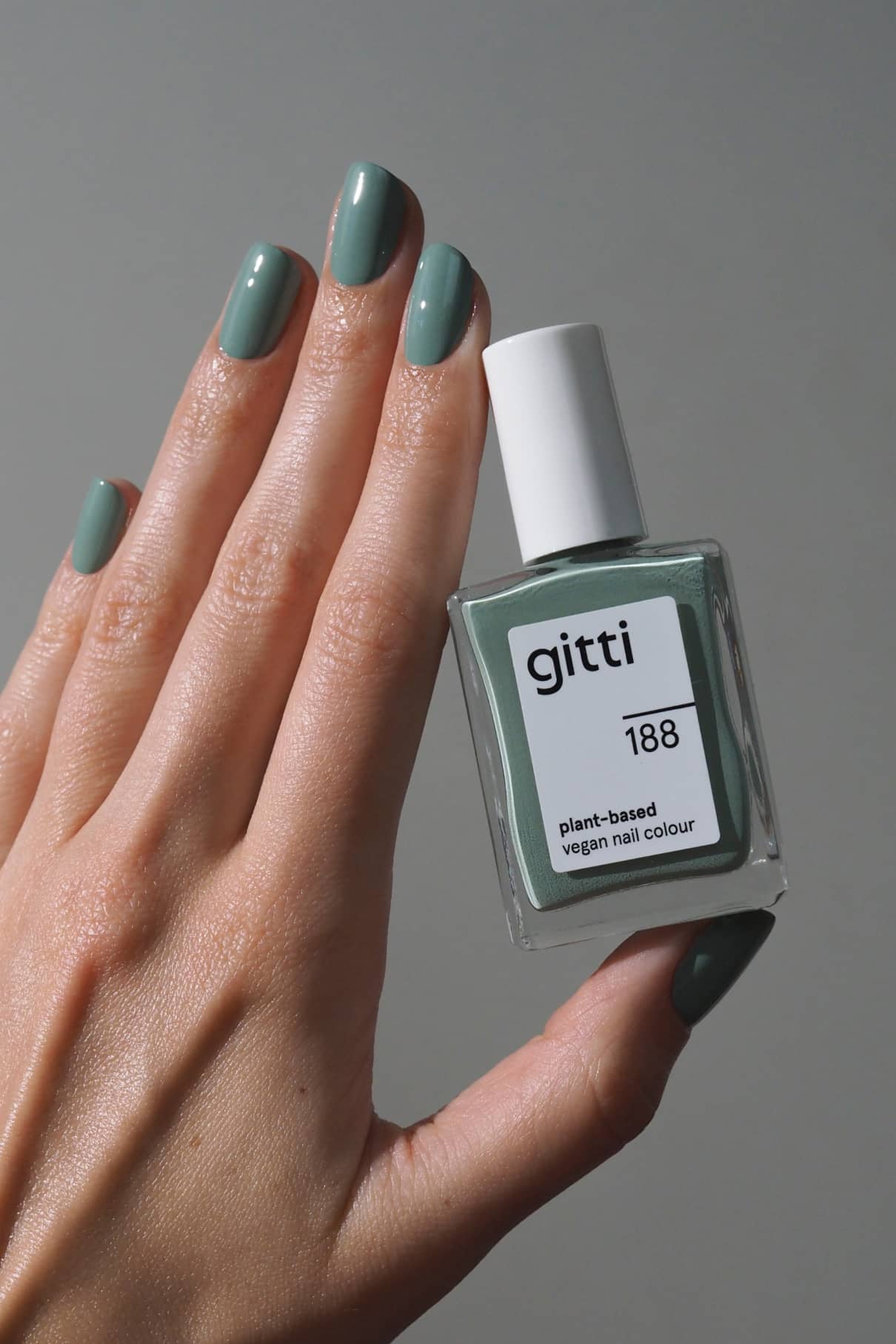 Eine Hand mit grün lackierten Nägeln hält ein Fläschchen mit gitti no. 188 pflanzlicher veganer Nagelfarbe vor einem schlichten grauen Hintergrund.