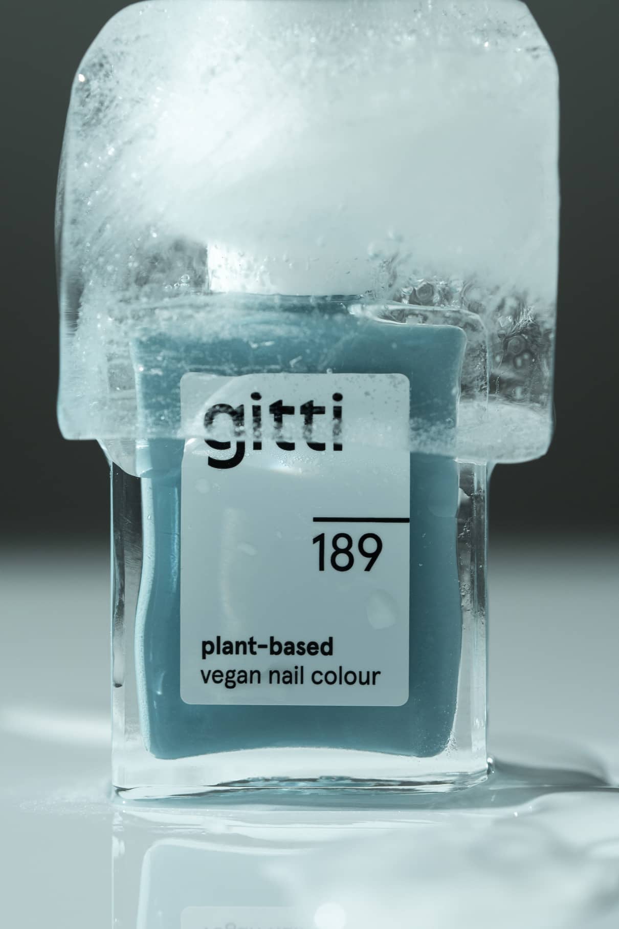 Ein Fläschchen von gitti no. 189 pflanzlicher veganer Nagellack in einem gedeckten Blauton ist teilweise von klarem Eis auf einer reflektierenden Oberfläche umhüllt.