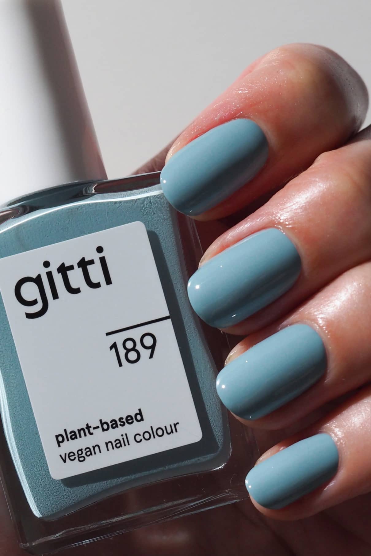 Eine Hand mit kurzen, sauber manikürten und in einem gedeckten Blauton lackierten Nägeln hält eine Flasche gitti no. 189 pflanzlichen veganen Nagellack im passenden Farbton.