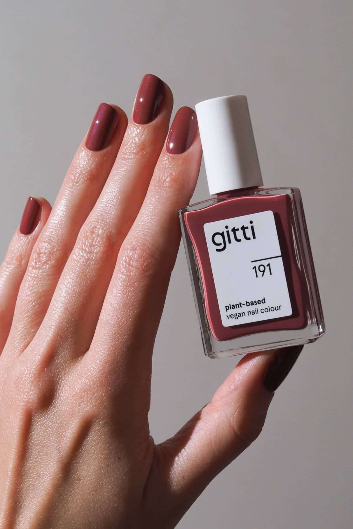 Eine Hand mit kurzen, glänzenden Beerenrosa-Nägeln hält eine Flasche gitti no. 191 pflanzlichen veganen Nagellack in einem passenden gedeckten Rot-Ton.