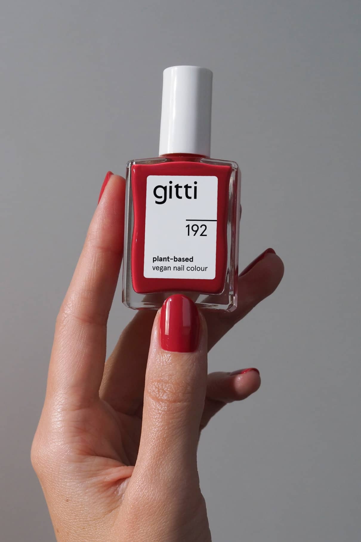 Eine Hand mit lackierten Nägeln hält eine Flasche von gitti no. 192, eine pflanzliche vegane Nagelfarbe in leuchtendes Rot, vor einem schlichten Hintergrund.
