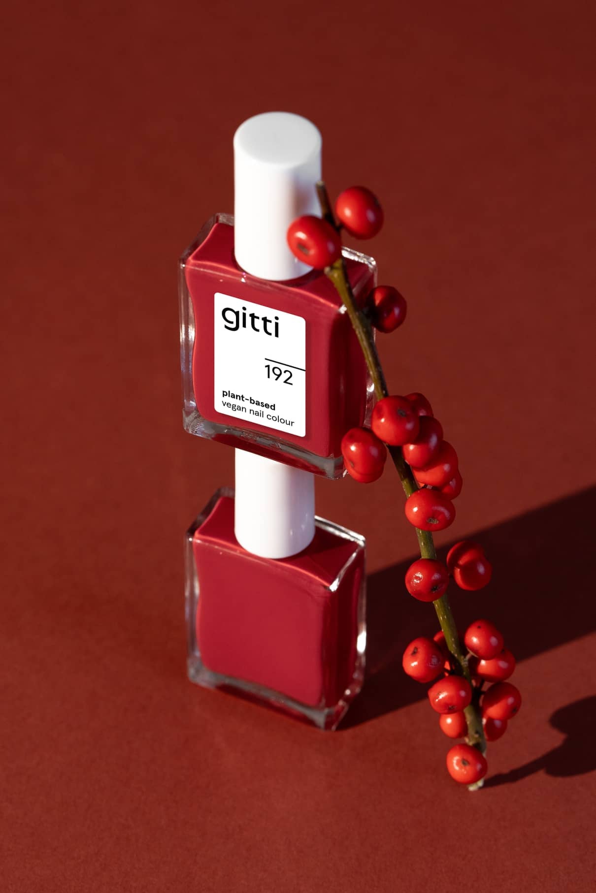 Zwei Flaschen von gitti no. 192 pflanzlicher veganer Nagellack in leuchtendes Rot sind senkrecht gestapelt, ein Zweig mit roten Beeren lehnt auf ihnen vor einem rotbraunen Hintergrund.