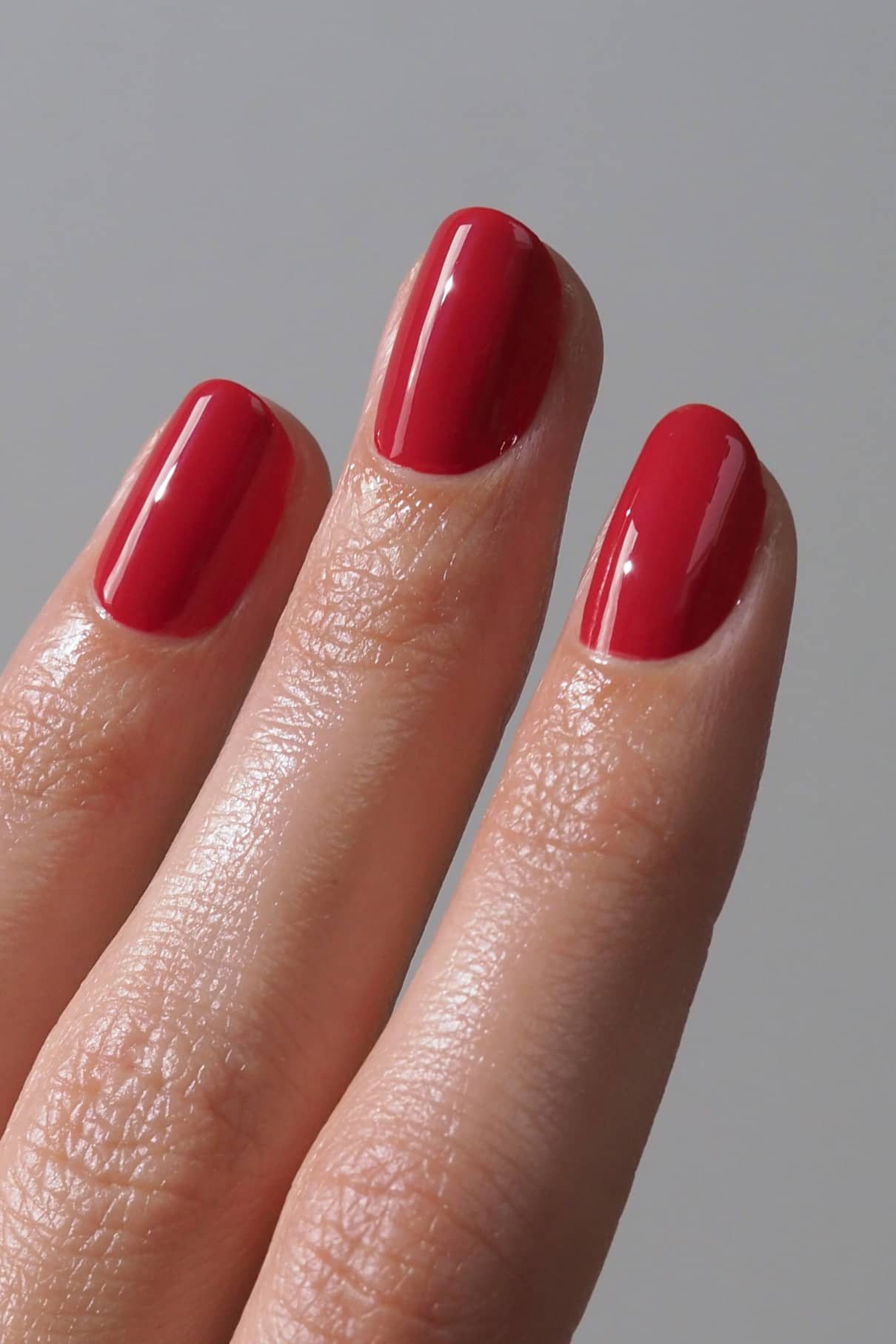 Drei Finger mit sauber manikürten Nägeln, lackiert mit gitti no. 192, einem glänzenden roten Lack, vor einem schlichten grauen Hintergrund.