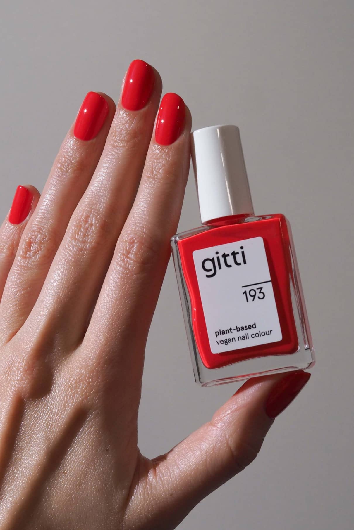 Eine Hand mit rot lackierten Nägeln hält ein Fläschchen mit gitti no. 193 pflanzlicher veganer Nagelfarbe in kräftigem Rot vor einem schlichten Hintergrund.