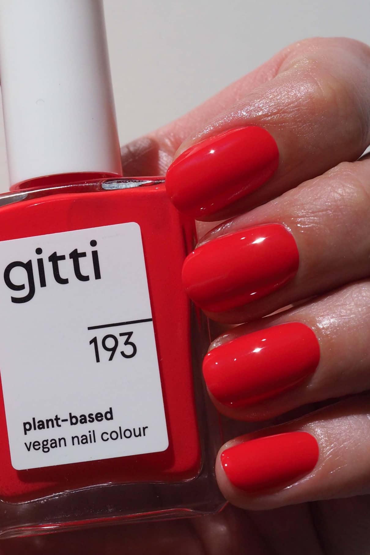 Eine Hand mit sauber manikürten, in einem leuchtenden Rot-Orange-Ton lackierten Nägeln hält eine Flasche gitti no. 193 pflanzlicher veganer Nagelfarbe.
