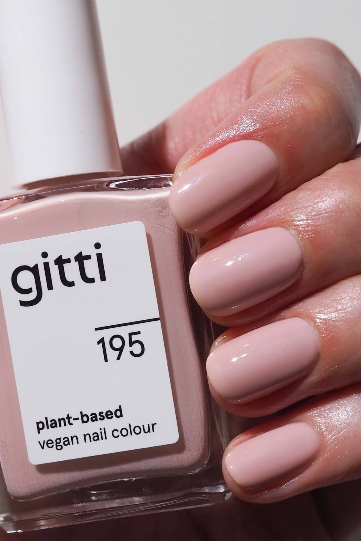 Eine Hand mit sauber manikürten Nägeln, die in einem pudrigen Rosé-Ton lackiert sind, hält eine Flasche mit gitti no. 195 veganen Nagellack auf Pflanzenbasis.